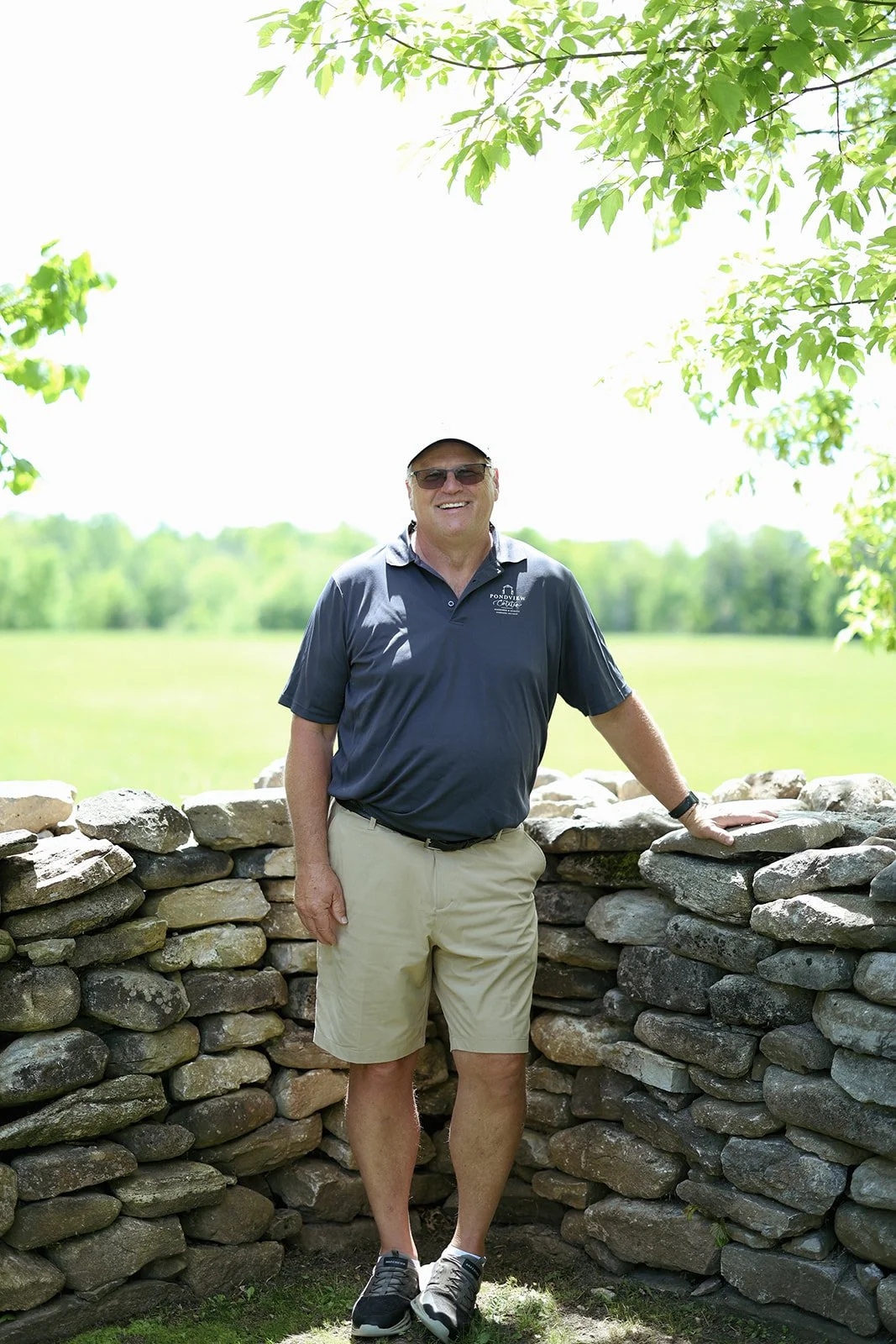 A middle-aged man wearing sunglasses, a navy blue polo shirt, beige shorts, and sneakers, standing outdoors with a smile. He is leaning on a stone wall with greenery and a field in the background.