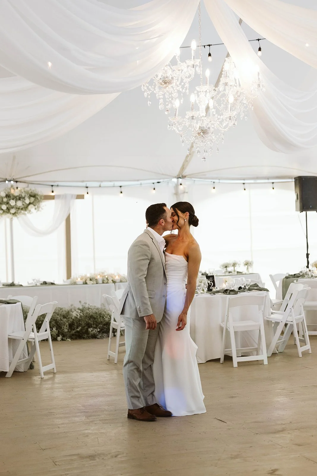 A bride and groom share a kiss under a decorated wedding tent with draped fabric and a chandelier.