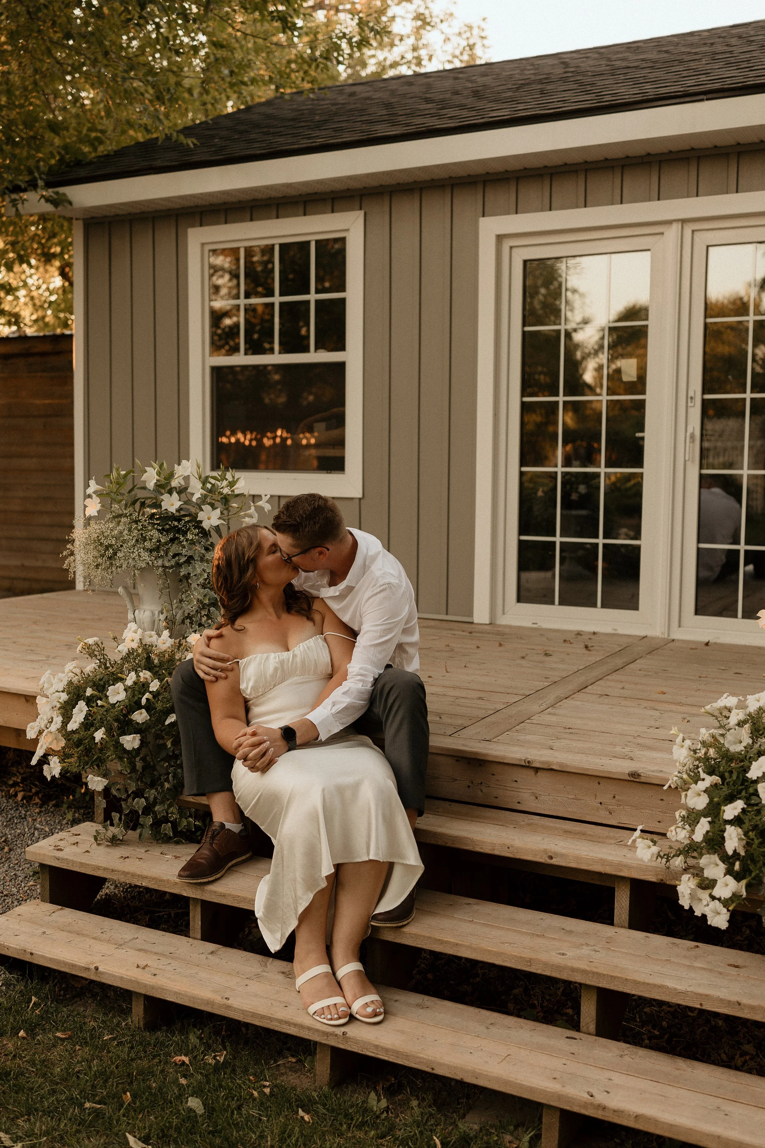 A couple kissing on wooden steps outside a house with a deck, surrounded by white flowers.