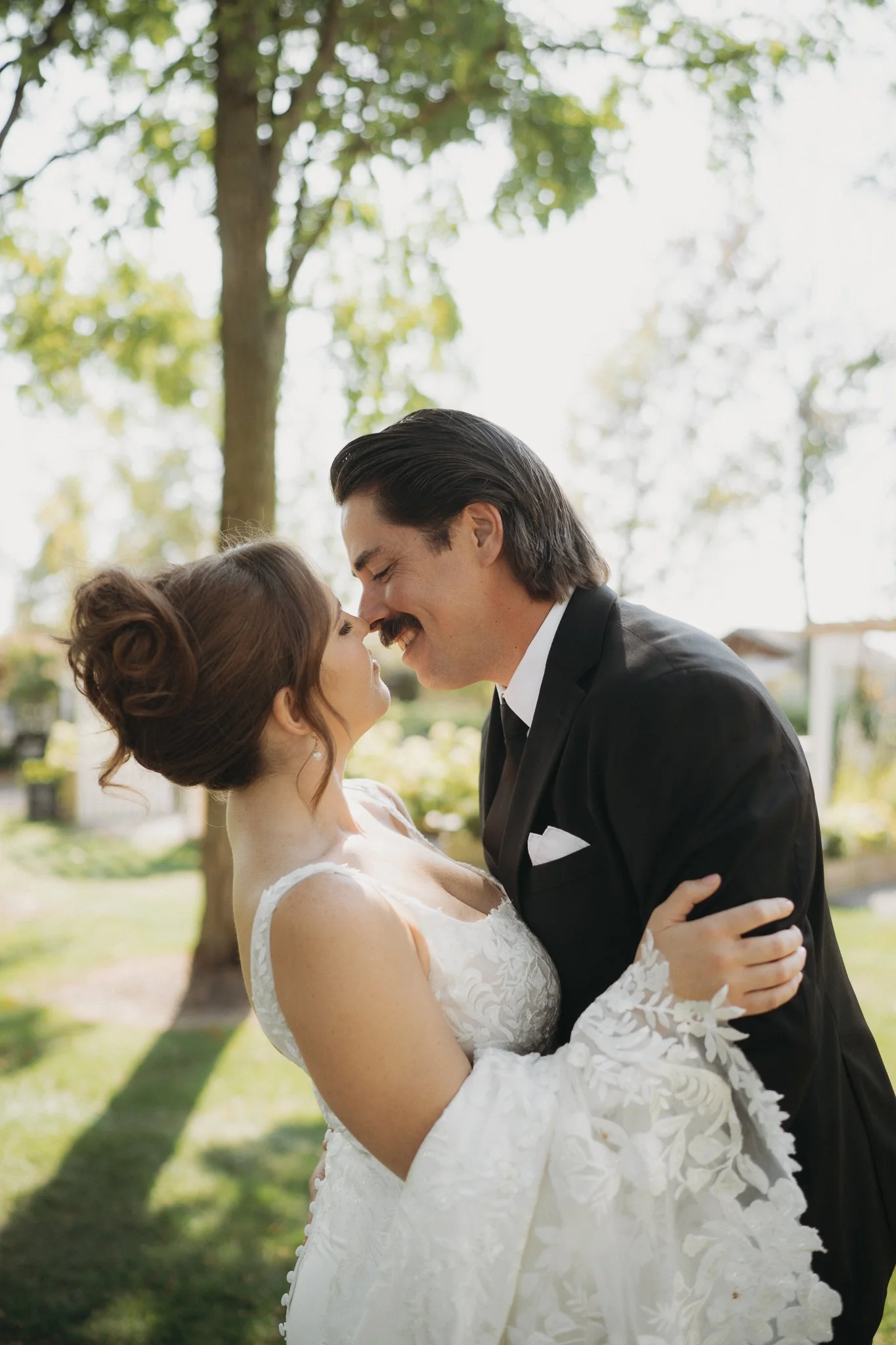 A bride and groom lovingly embrace outdoors, touching their foreheads together with eyes closed, in a sunny garden with trees in the background.