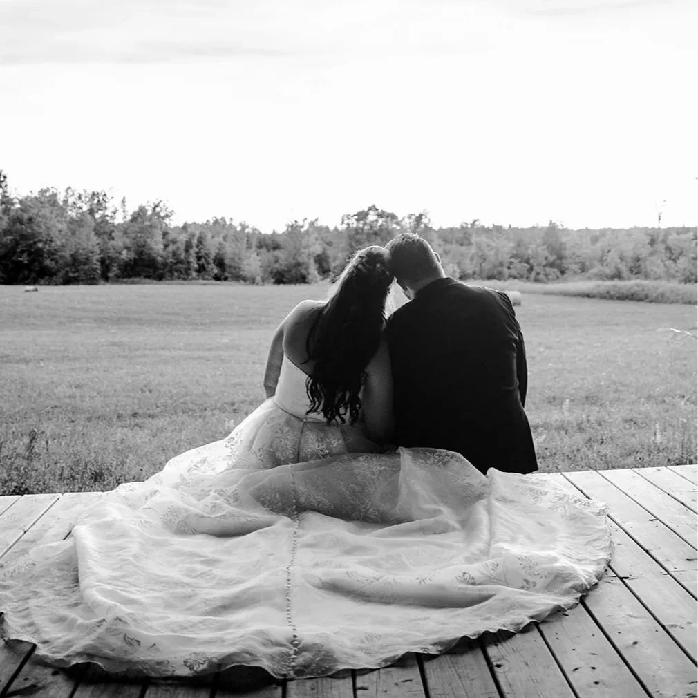 A couple sitting on a wooden porch, the woman in a wedding dress and the man in a suit, looking at a scenic outdoor landscape.