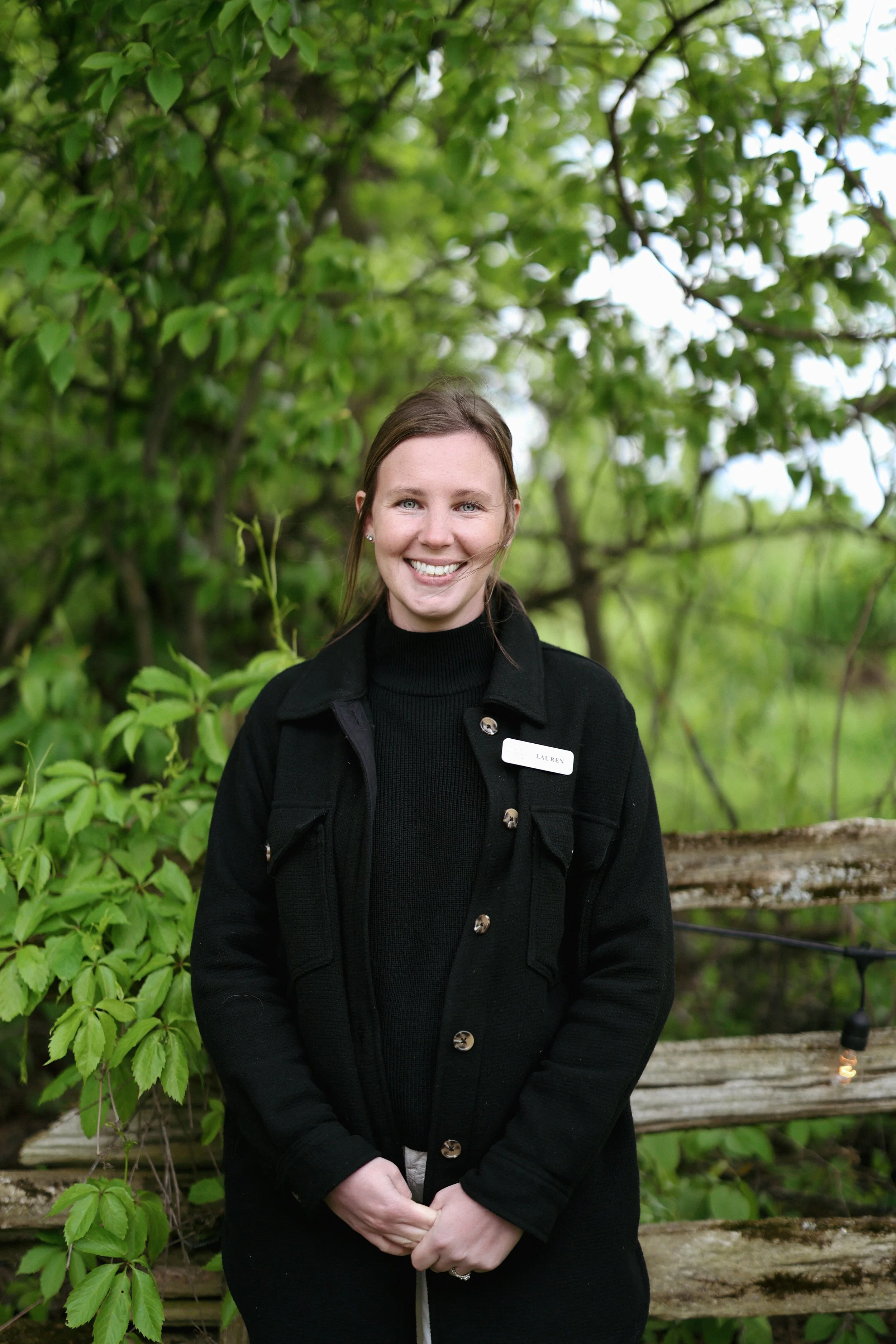 A woman smiling outdoors near green foliage and trees.
