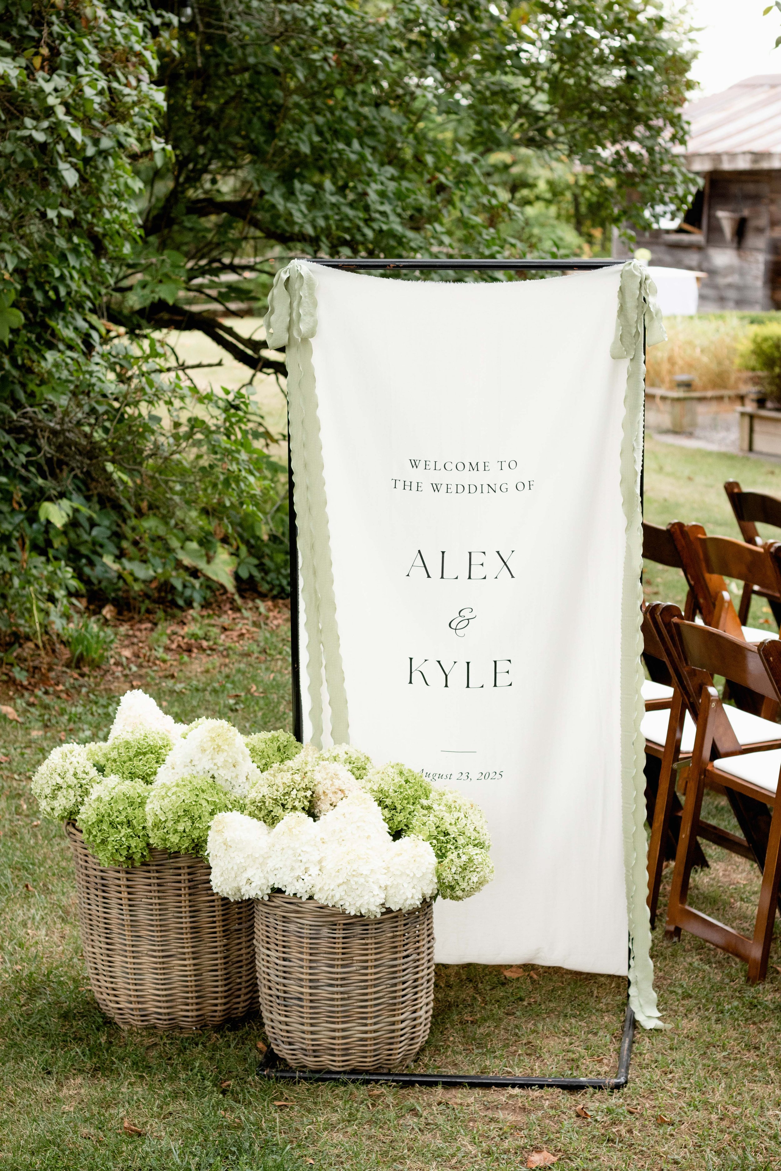 Outdoor wedding sign welcoming guests to Alex and Kyle's wedding on August 23, 2025, with white flowers in woven baskets nearby.