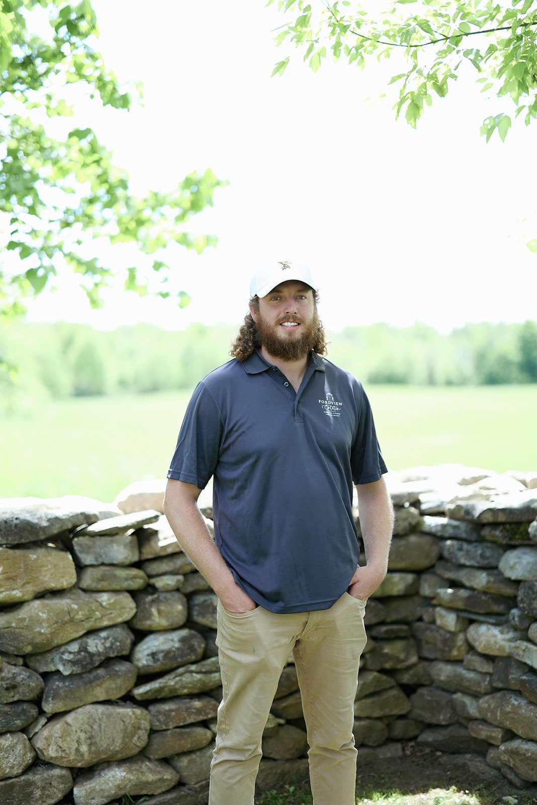 A young man with curly hair, a beard, wearing a white cap and dark blue polo shirt, standing outdoors in front of a stone wall, with green trees and a grassy field in the background.