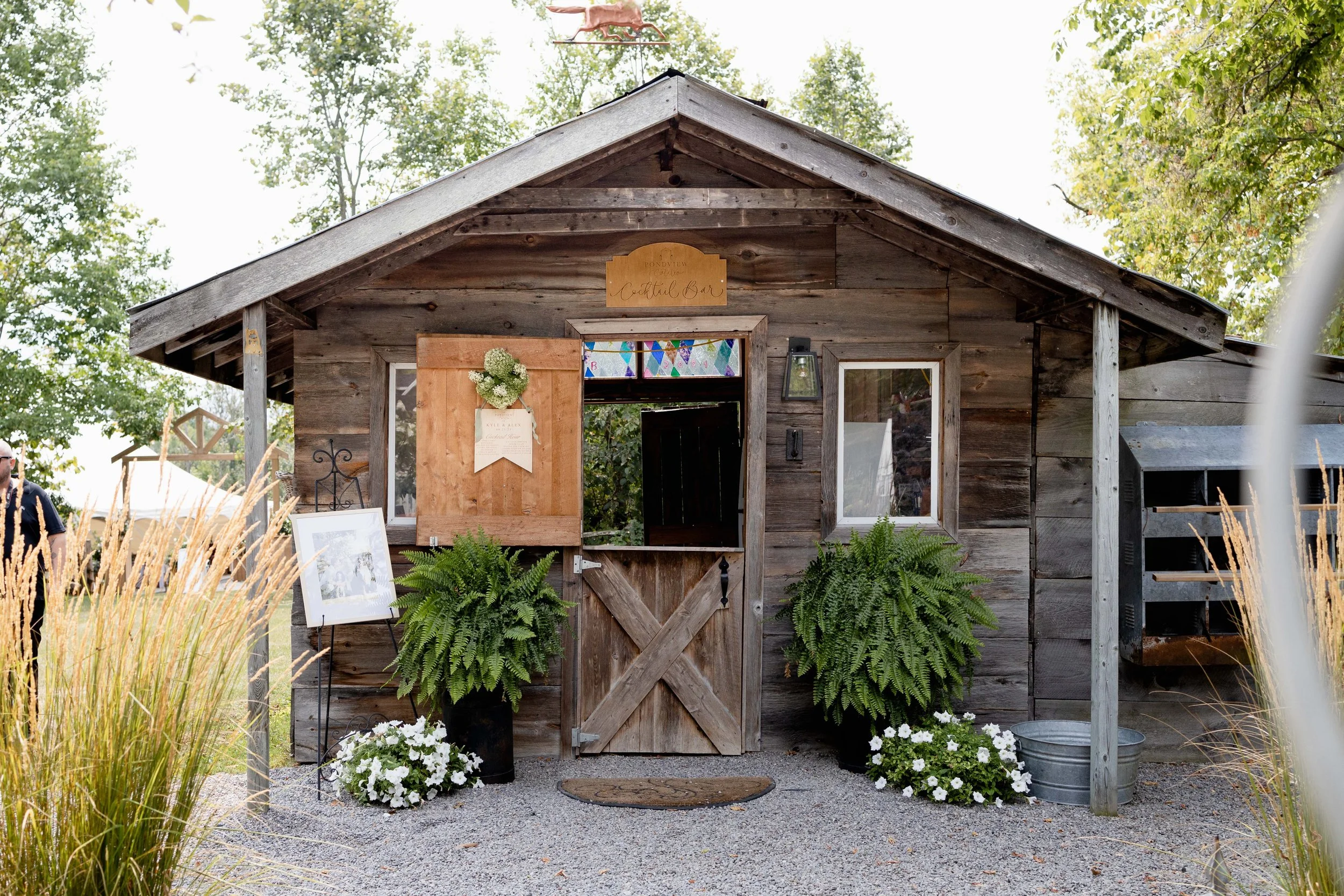 Small rustic wooden building with a sign that says 'Cocktail Bar' above the door, decorated with potted ferns and white flowers, and framed photographs outside.