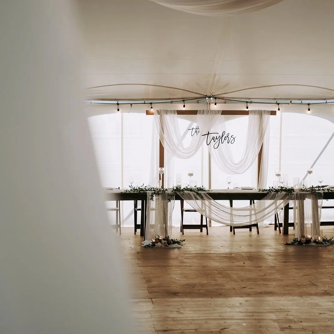 Indoor wedding reception setup with a long table draped in white fabric, decorated with greenery and flowers, positioned in front of large windows with sheer curtains and a sign that says 'the Taylors'.