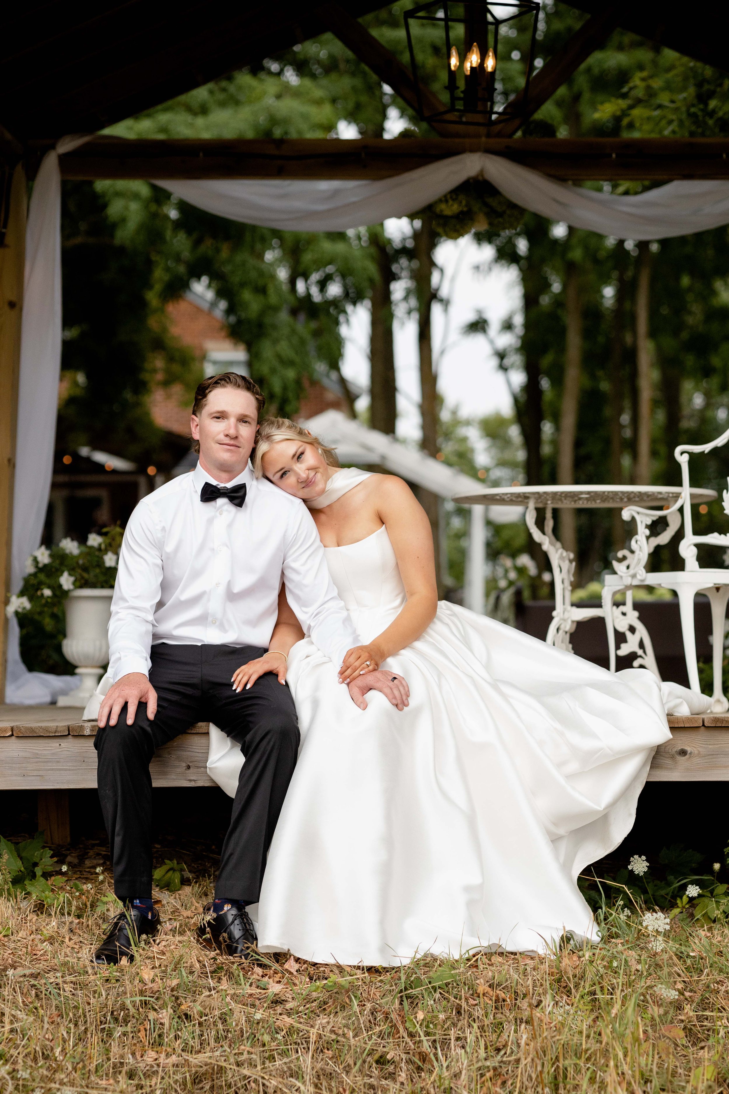 A newlywed couple sitting on a wooden platform outdoors, with the bride resting her head on the bridegroom's shoulder, both dressed in wedding attire. The bride wears a white wedding gown and the groom wears a black tuxedo with a bowtie. There are trees in the background and decorative elements under a shelter above them.