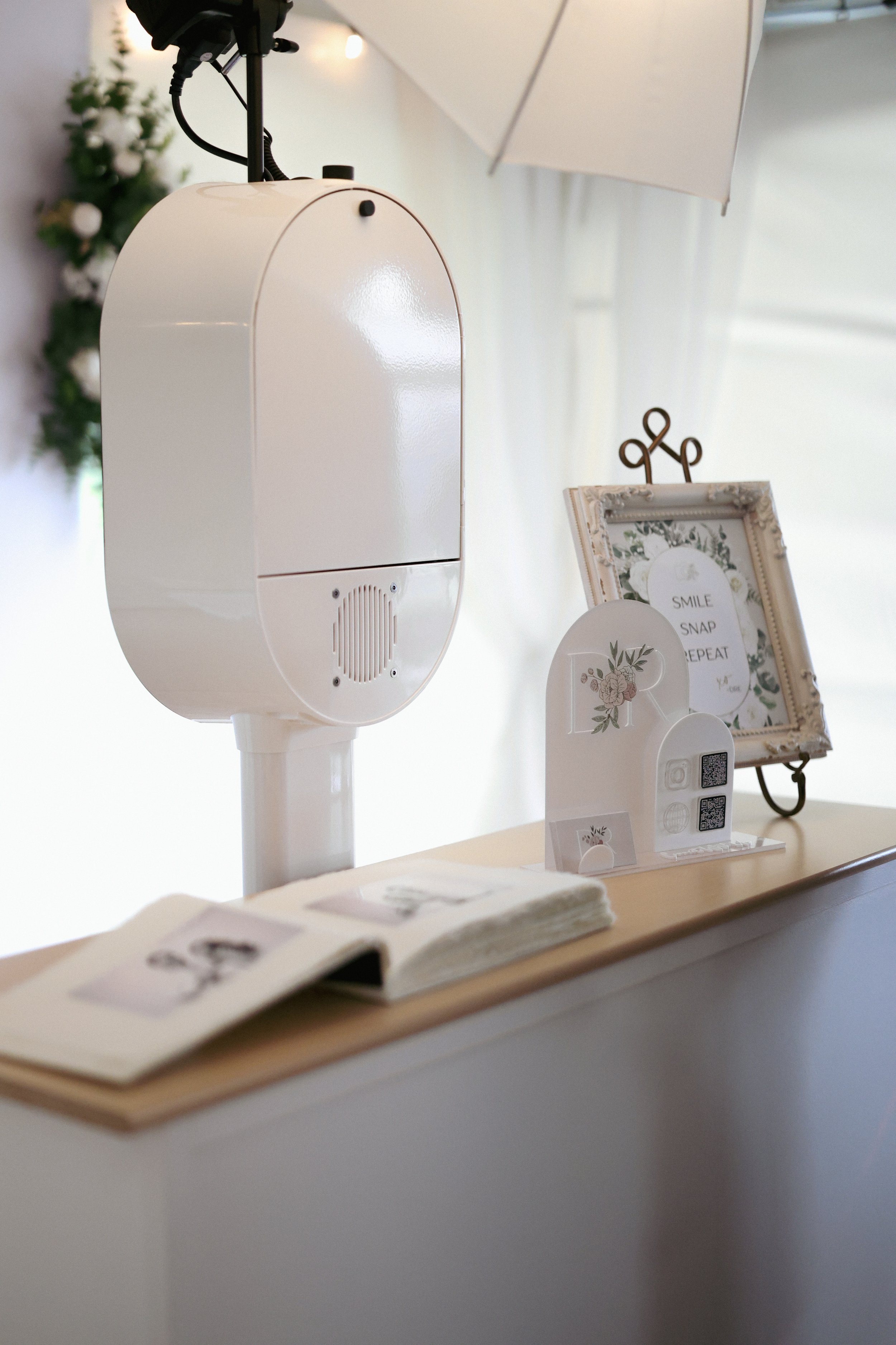 White photo booth with camera on stand, framed sign with floral design and the words 'Smile, Snap, Repeat', and floral-themed decorations on a table.
