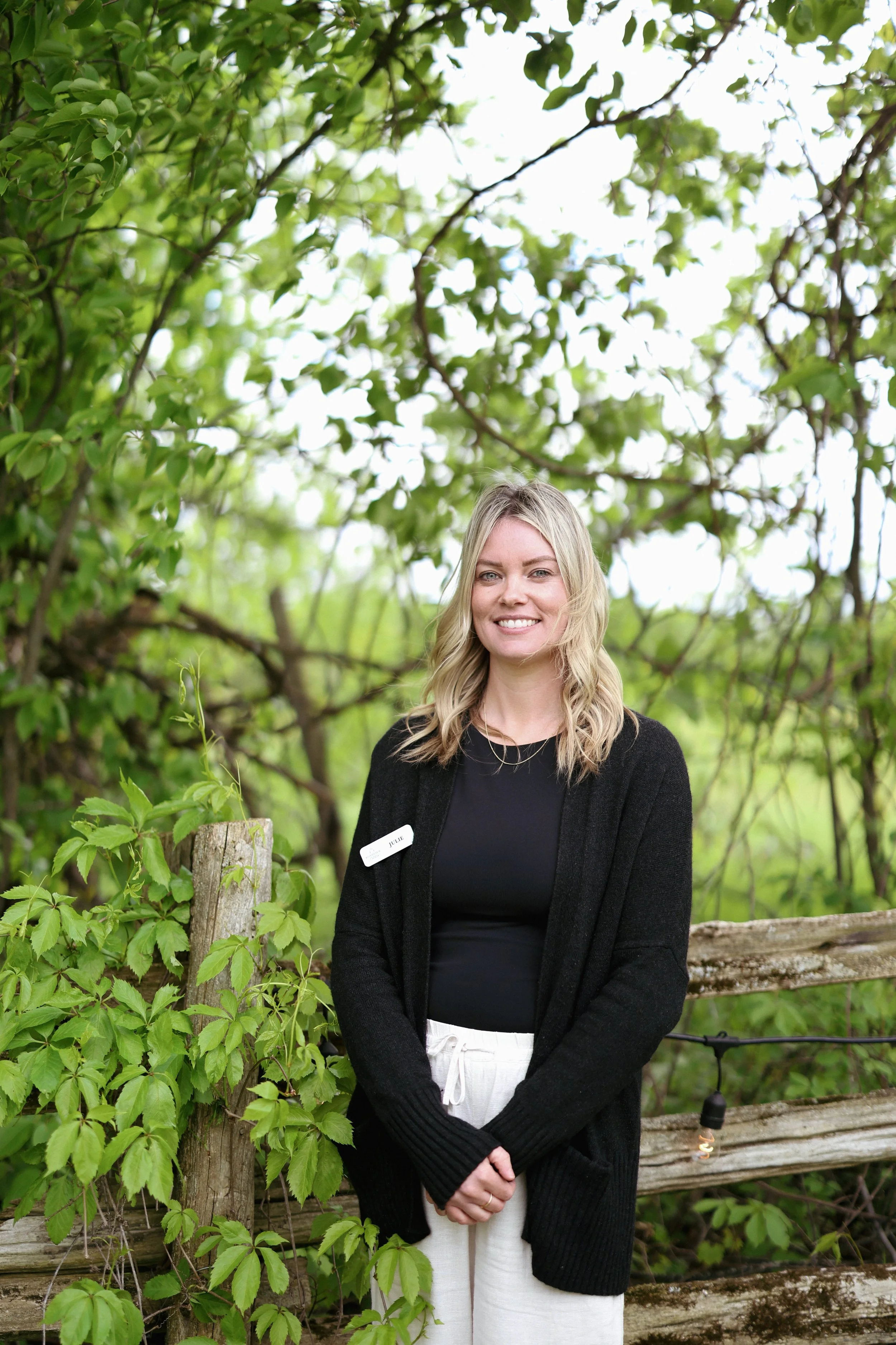 A smiling woman standing outdoors near a rustic wooden fence and green foliage.