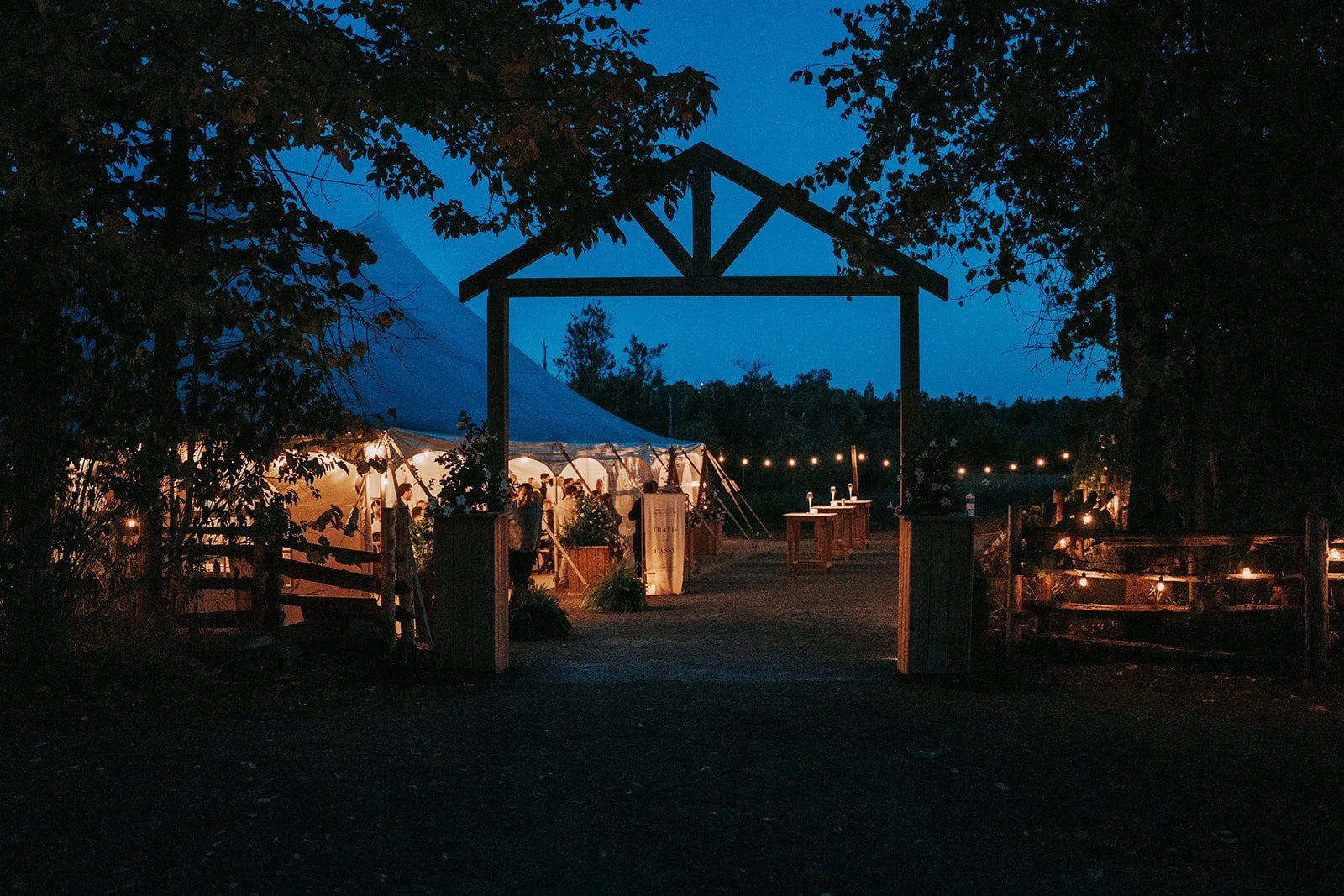 A nighttime outdoor event area with a large white tent, string lights, and wooden tables, viewed through an open wooden gate with trees surrounding the scene.
