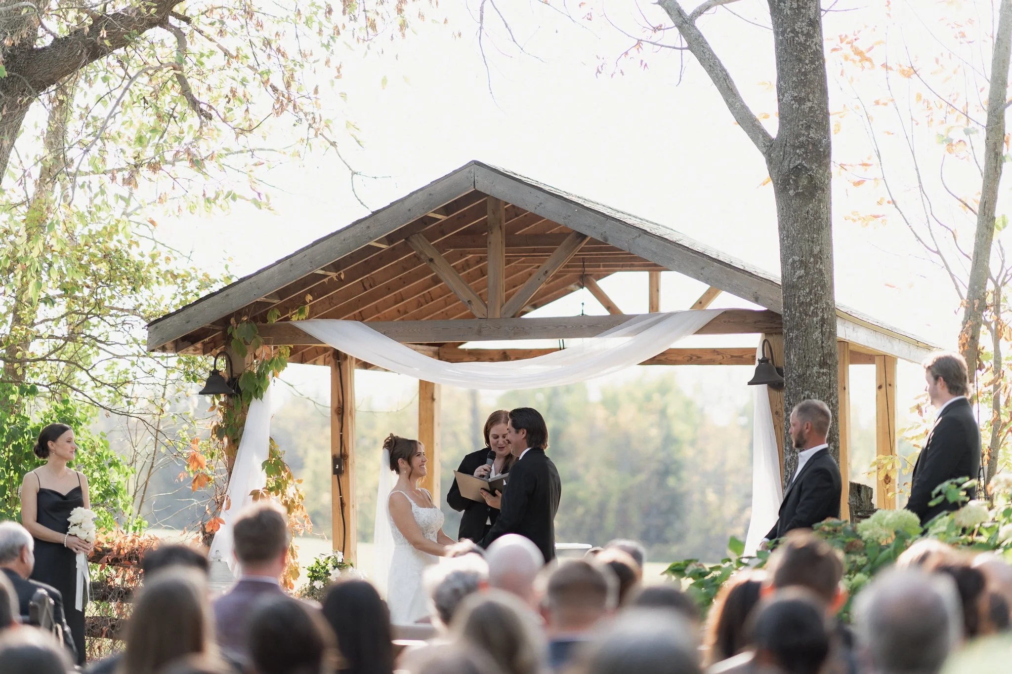 A wedding ceremony takes place outdoors under a wooden pavilion with a triangular roof. The bride and groom are holding hands and facing each other, with a woman officiating over the ceremony. Guests are seated facing them. The scene is surrounded by trees with green and some autumn-colored leaves, with sunlight filtering through.