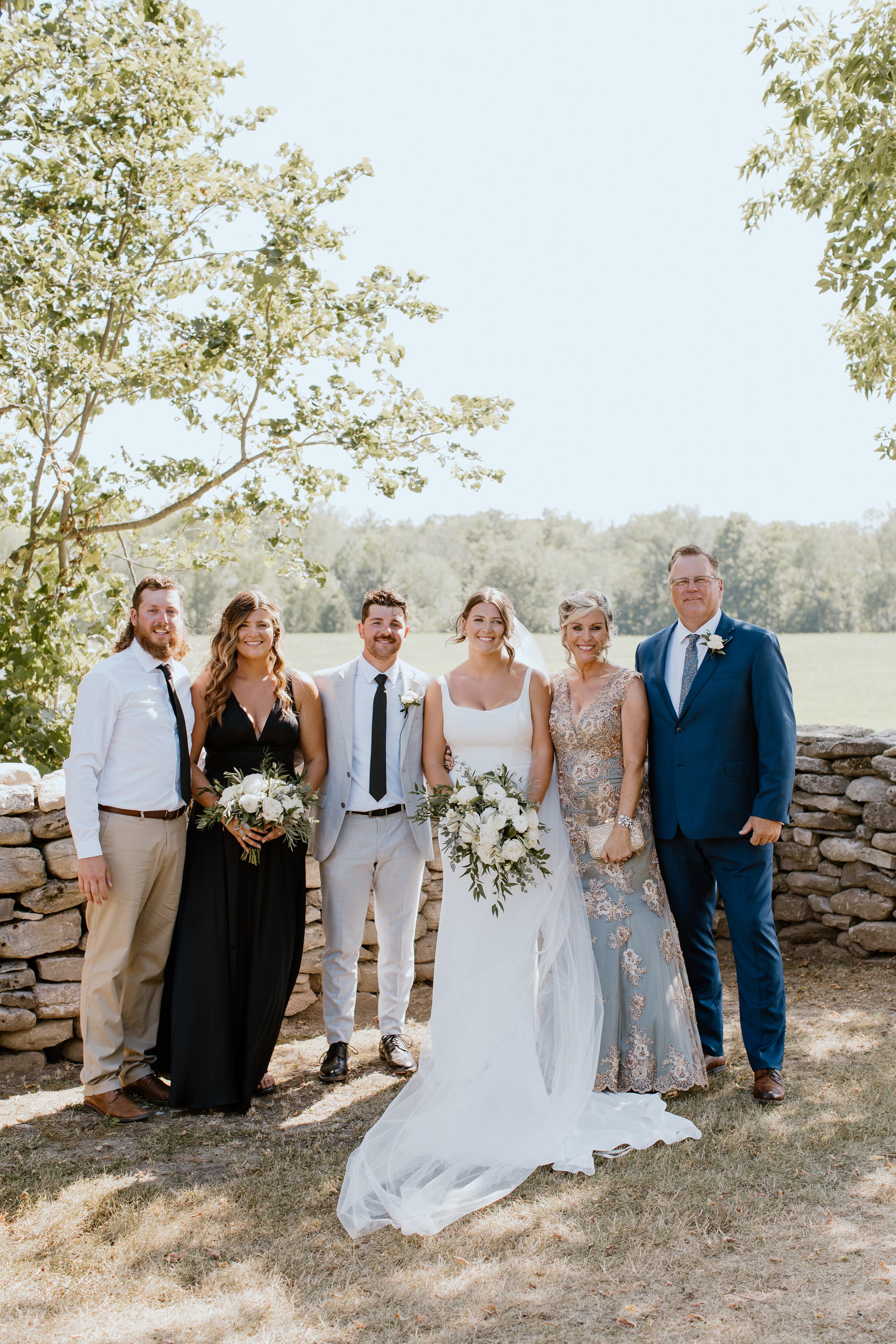 A wedding party standing outdoors in front of a stone wall with trees and grassy field in the background. The bride is in the center wearing a white wedding dress and holding a bouquet. The groom and other guests are dressed in formal attire.