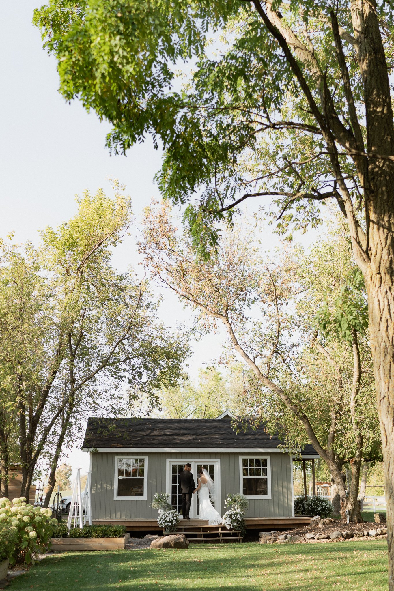 A couple in wedding attire holding hands and facing each other on the steps in front of a small, gray house surrounded by trees and greenery during daytime.