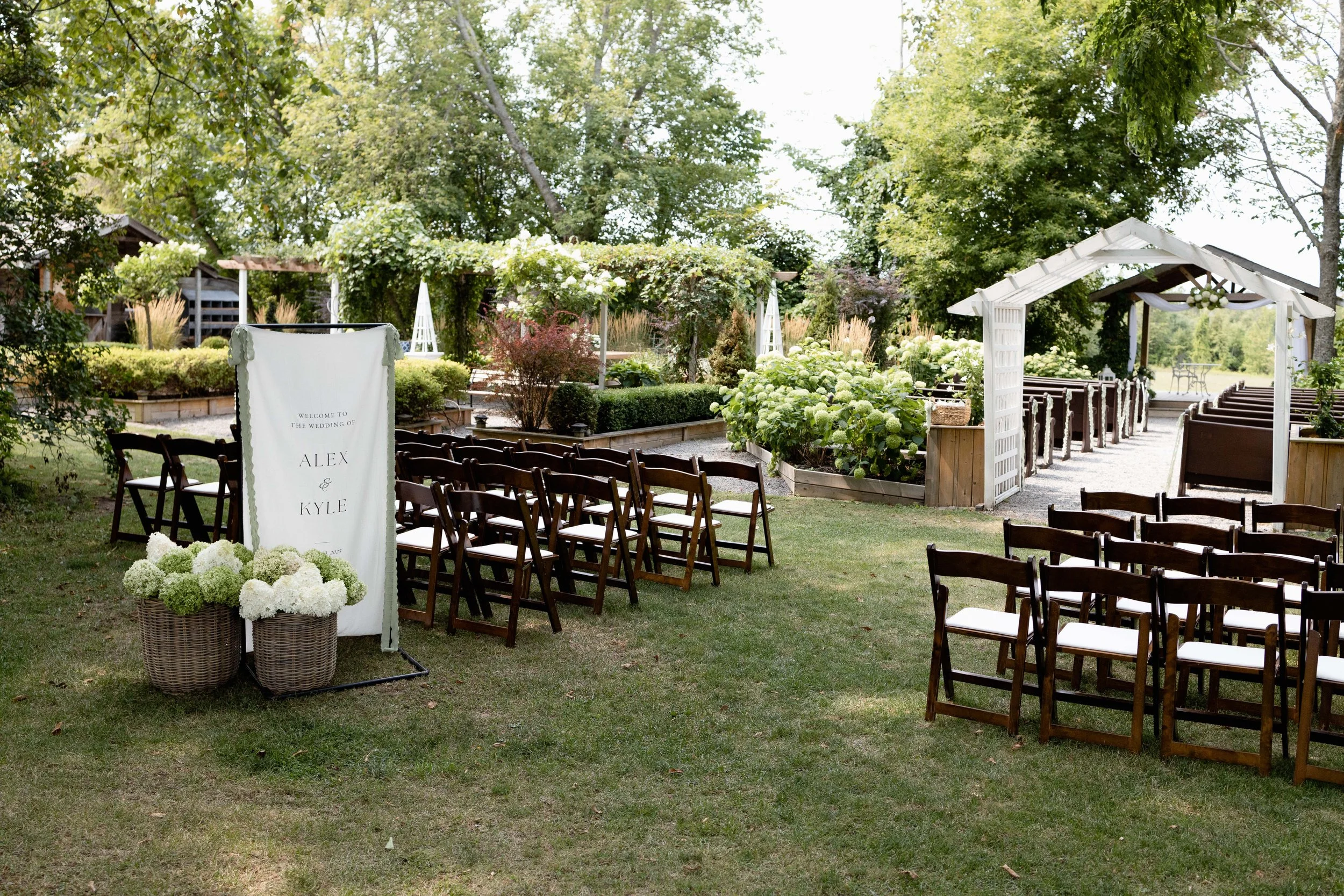 Outdoor wedding ceremony setup with rows of wooden chairs, a white arch at the front, and a sign that reads "Welcome to the wedding of Alex & Kyle." Flowers and greenery decorate the area, which is surrounded by trees and lush plants.