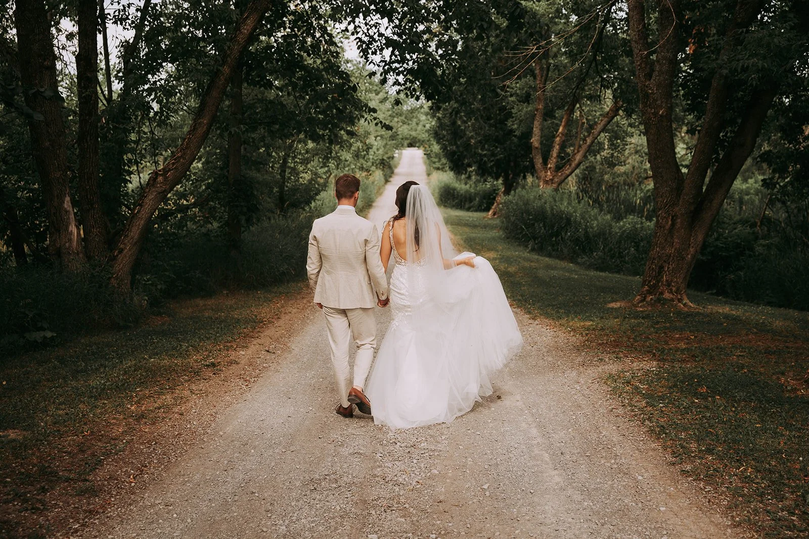 Bride and groom walking hand in hand on a dirt path through a wooded area, dressed in wedding attire, as seen from behind.