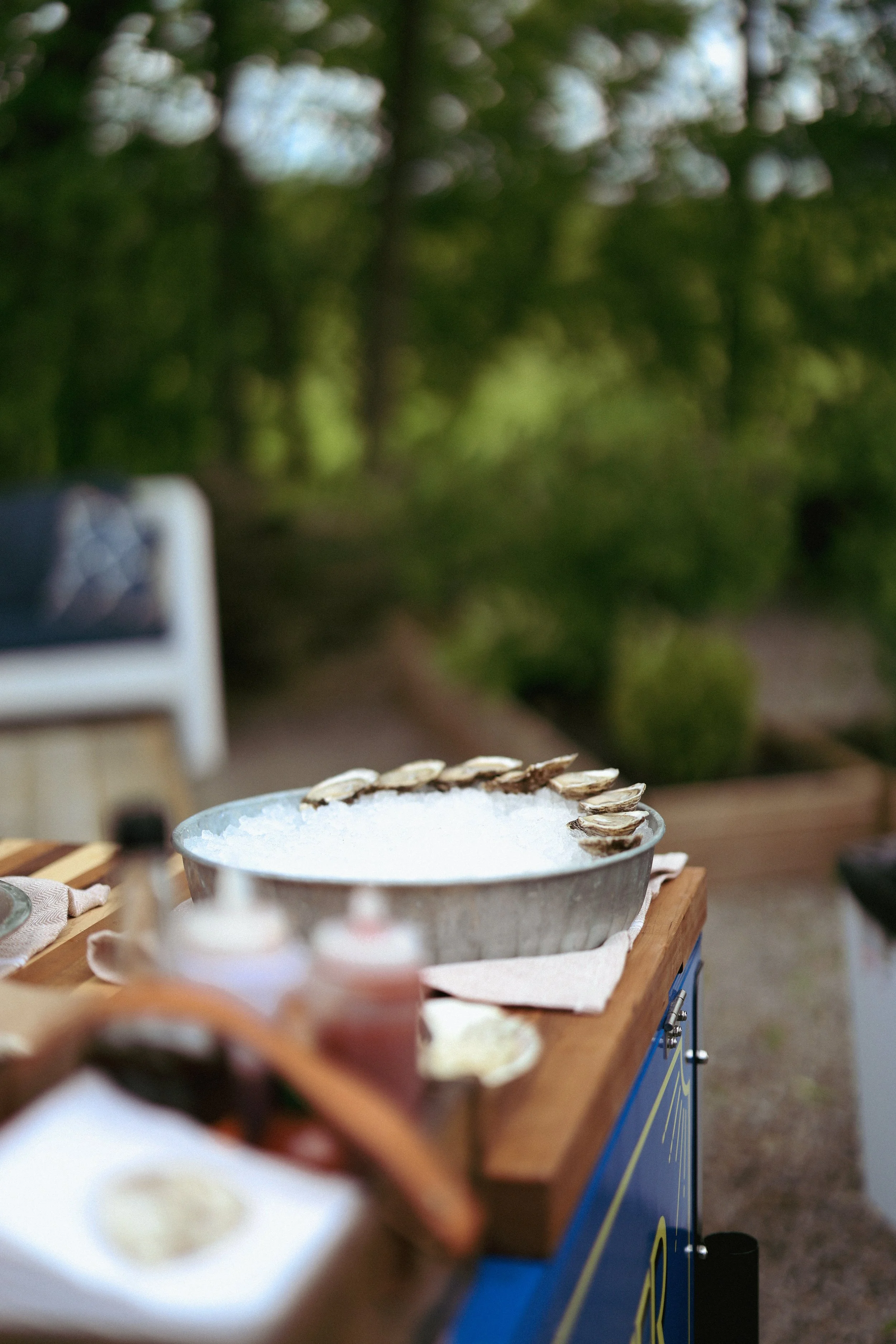 A metal bowl filled with frothy drink, garnished with clam shells, sitting on a wooden table outdoors in a green, wooded area.