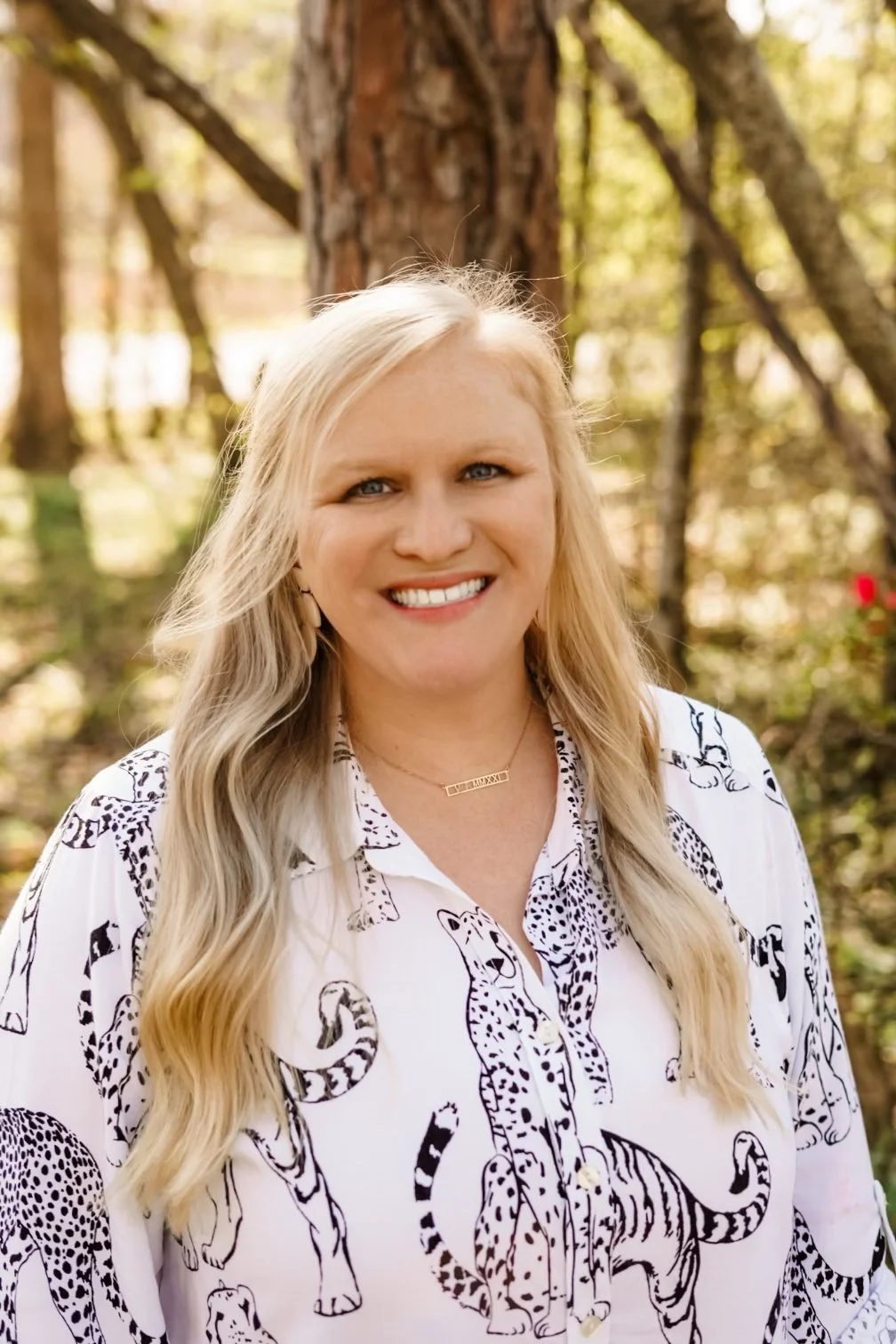 A woman with long blonde hair smiling outdoors in a wooded area, wearing a white shirt with black animal print and a gold necklace.
