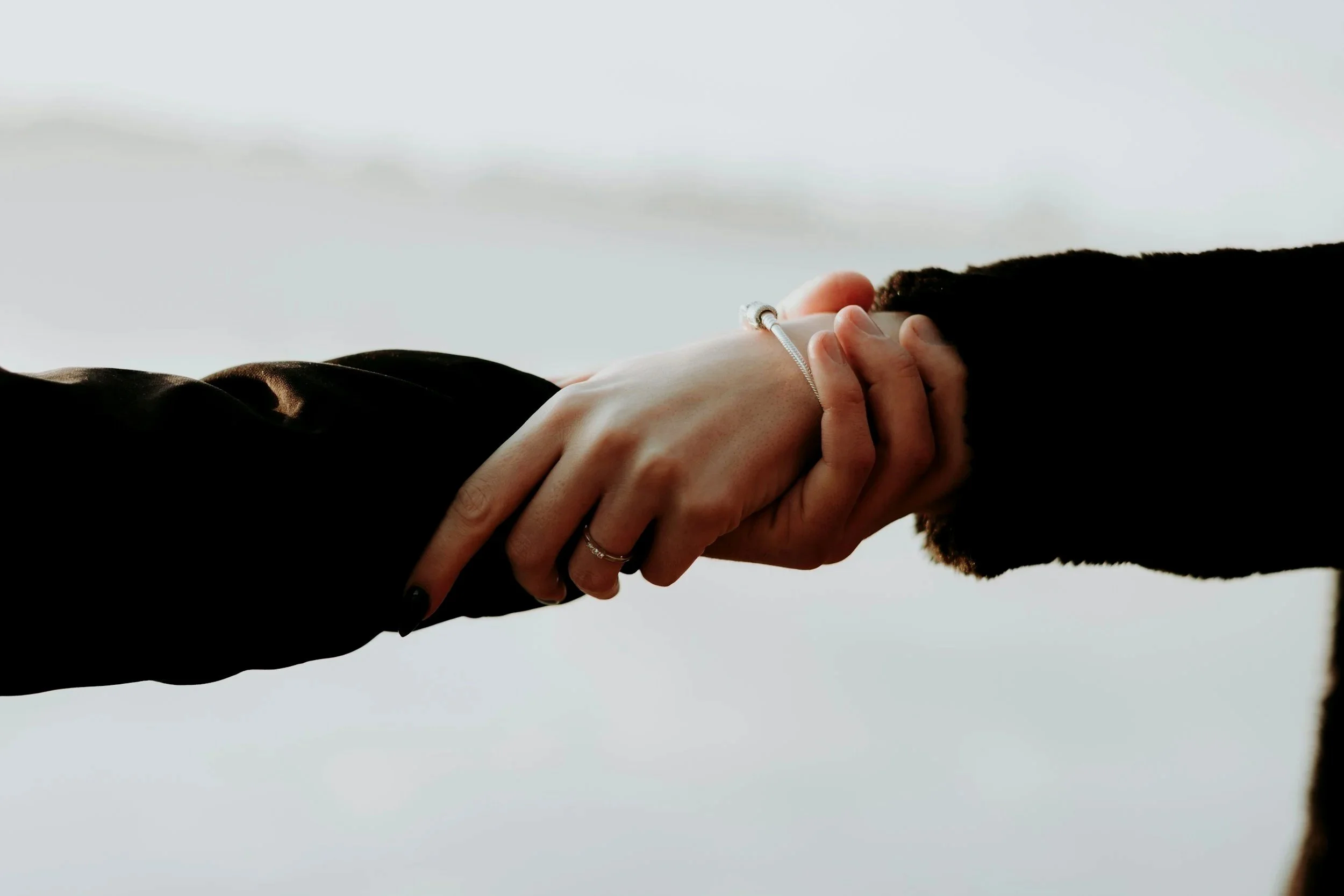 Two people holding hands, wearing black sleeves and jewelry, with a cloudy sky background.