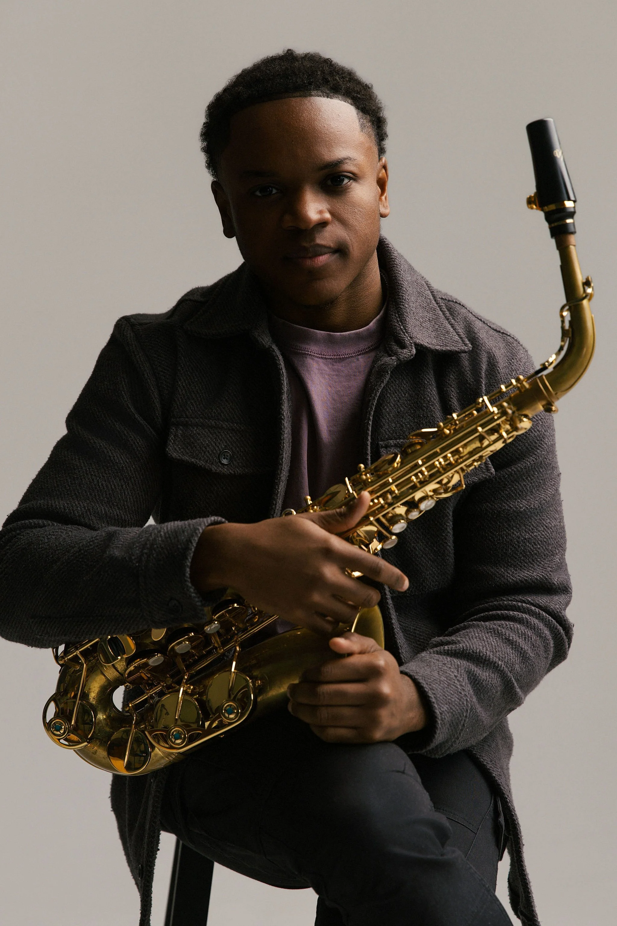 Langston Hughes II; A young man with short curly hair holding a gold saxophone while sitting on a stool against a plain background.