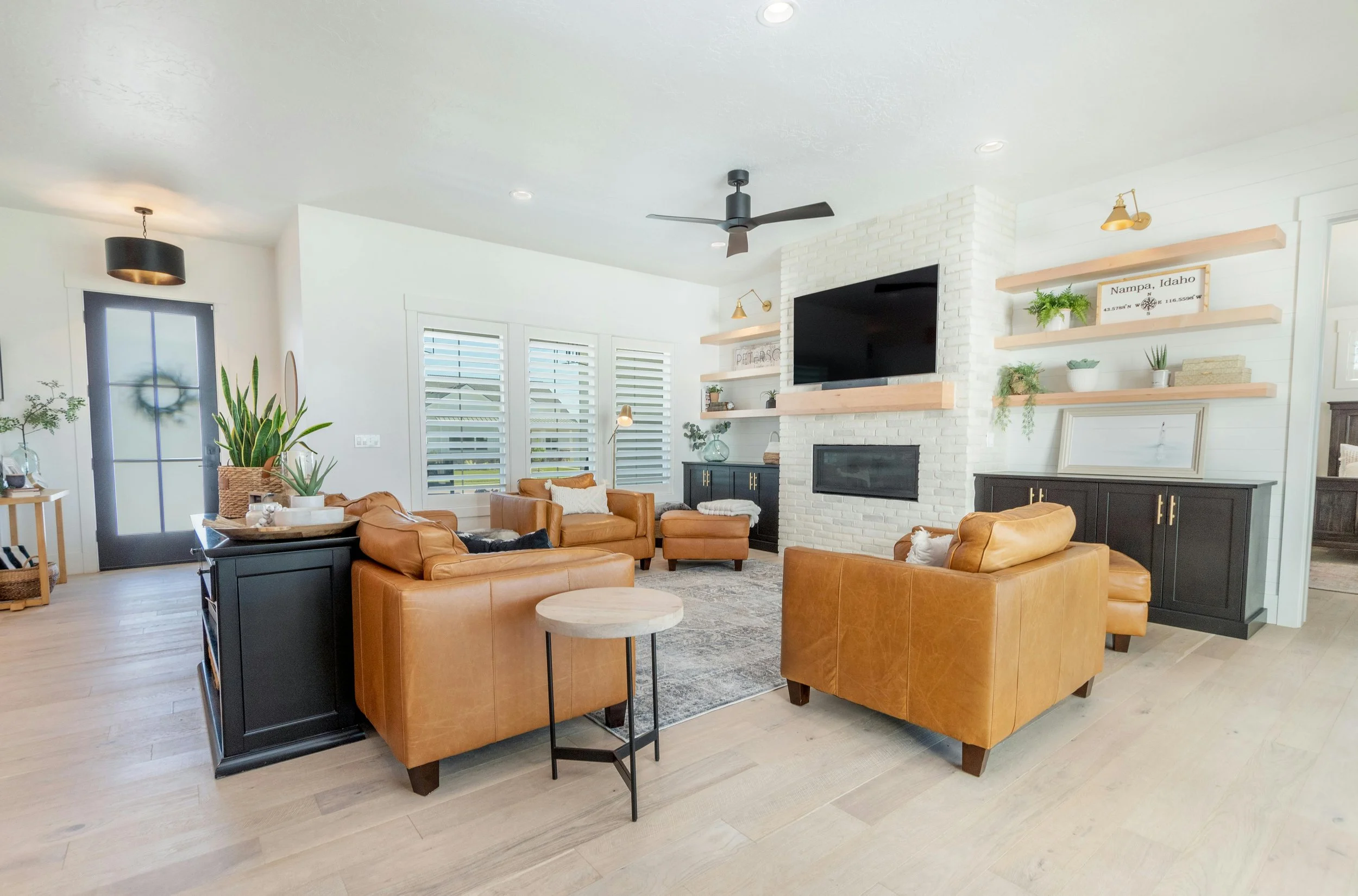 Living room with tan leather sofas, black cabinetry, white brick fireplace, mounted TV, wooden shelves with plants and decor, windows with white shutters, light wood flooring, and modern decor.