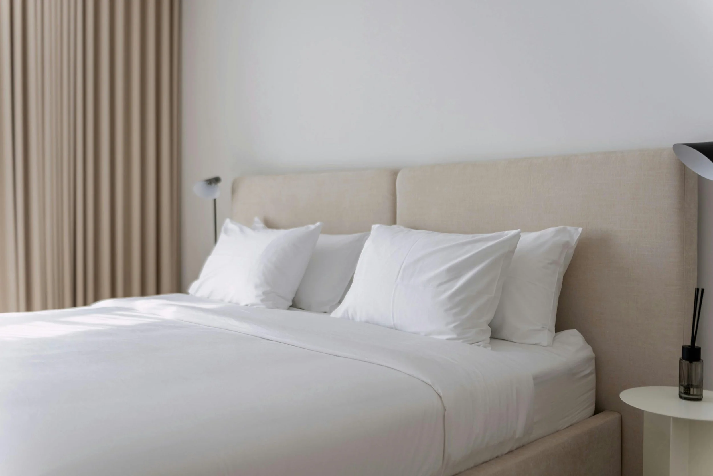 A neatly made bed with white linens and multiple white pillows in a modern bedroom. There is a beige headboard, beige curtains on the window, a white bedside table with a reed diffuser, and a wall-mounted reading lamp on each side of the bed.