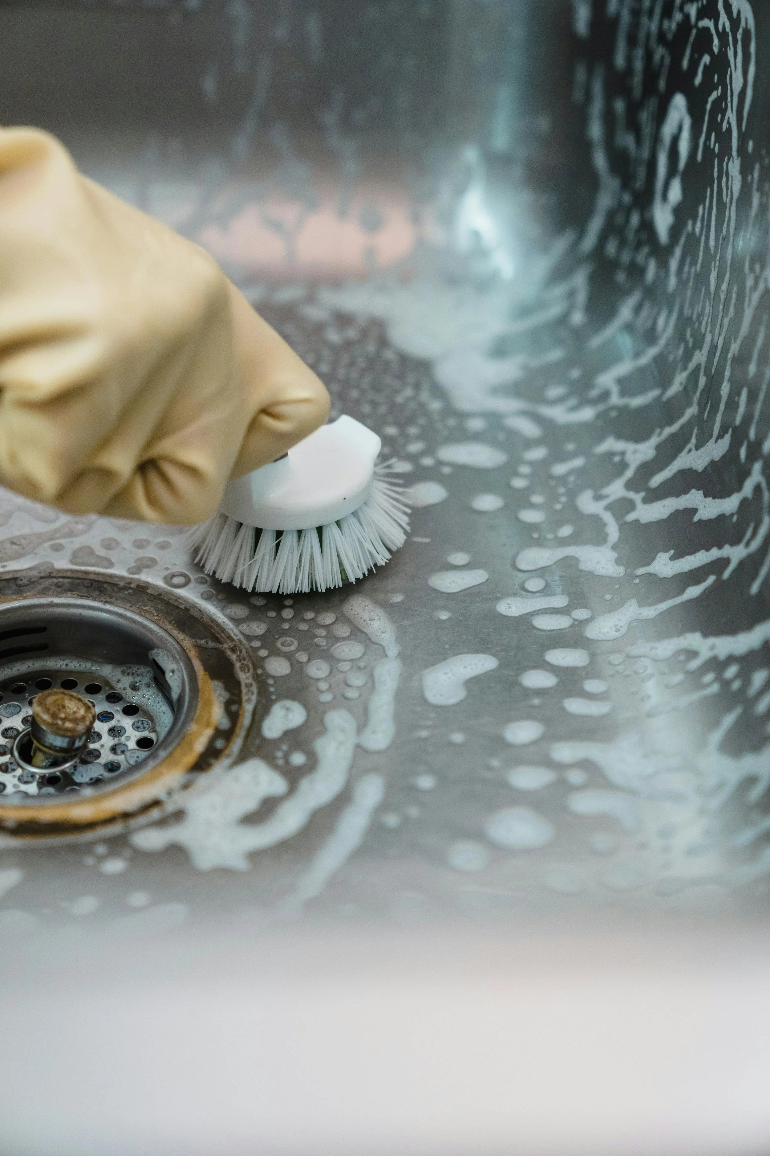 A gloved hand cleaning a stainless steel sink with a scrub brush and soap suds.