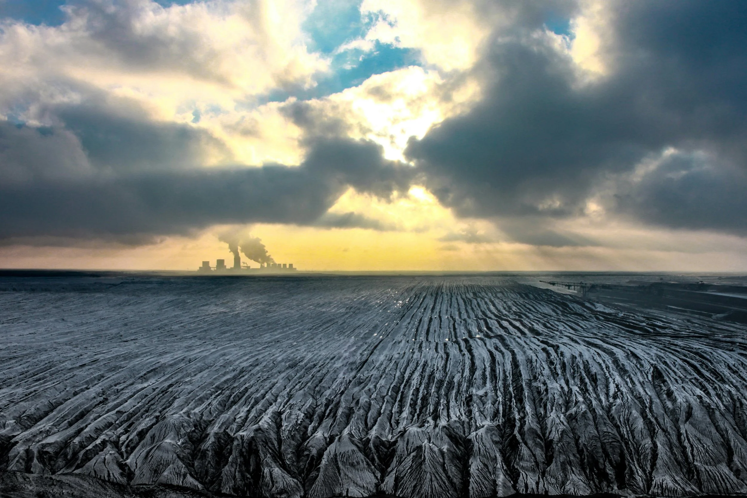 A vast, snowy landscape with plowed fields and dark tire tracks leading towards distant industrial buildings emitting smoke under a cloudy sky with sunlight breaking through.