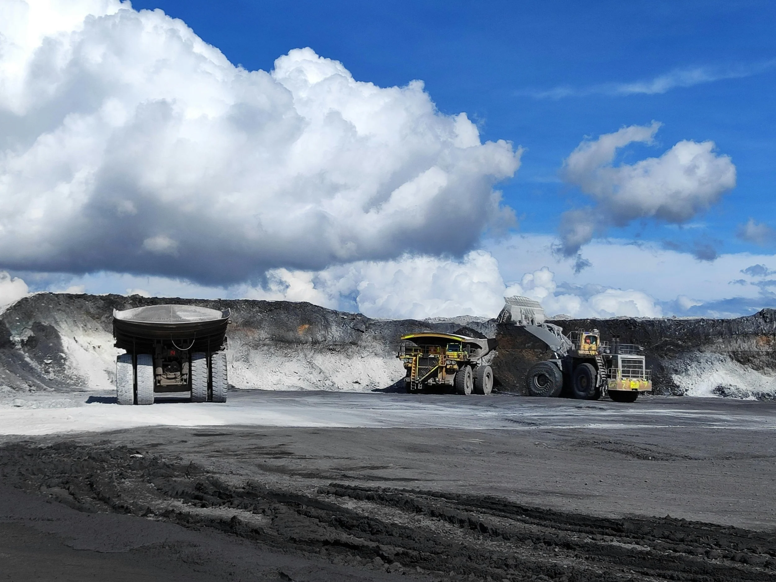 Large mining trucks operating on a rugged terrain with a partly cloudy sky overhead.