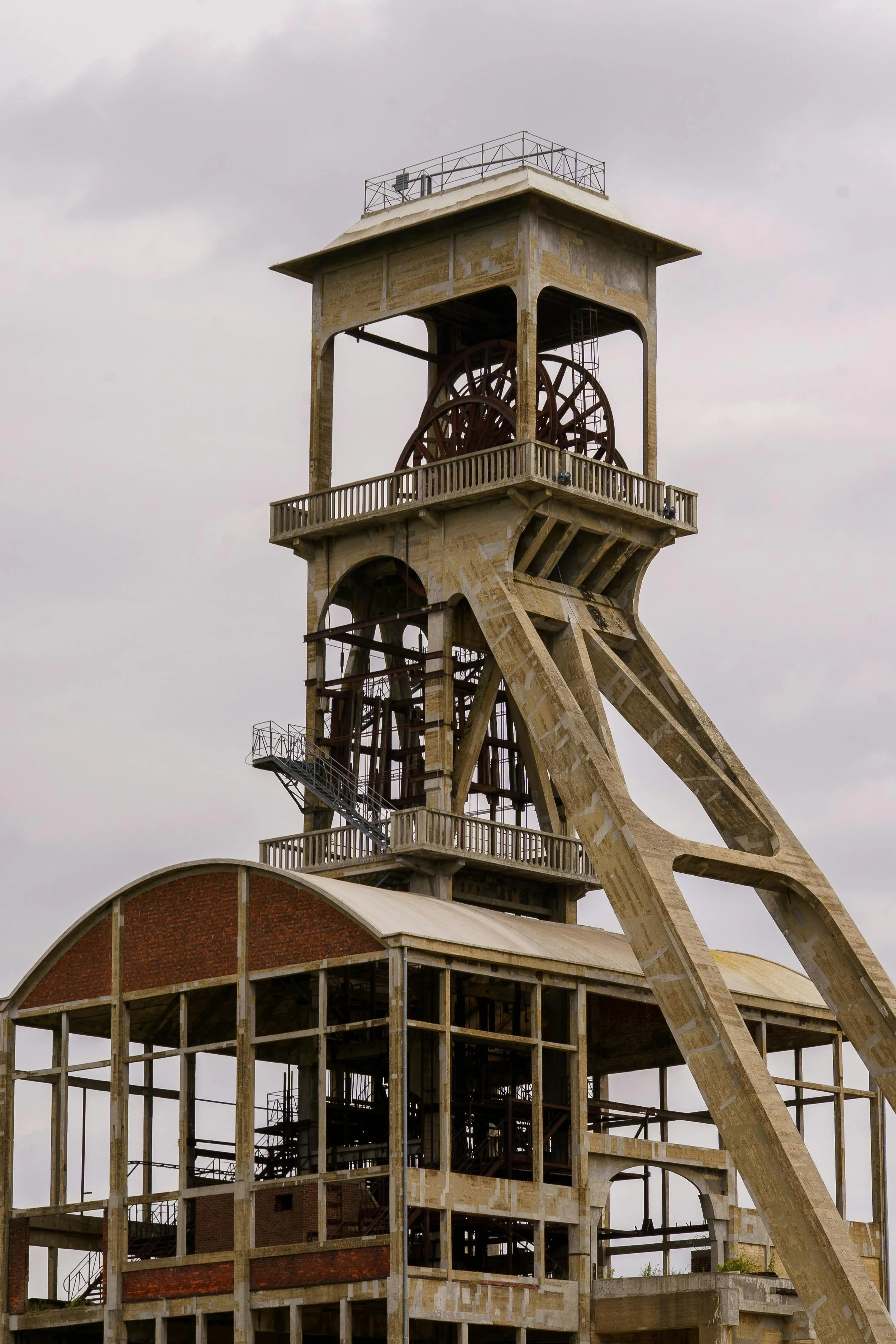 An old, abandoned mining headframe structure made of concrete and metal, with multiple levels, a large wheel, and open framework, under a cloudy sky.
