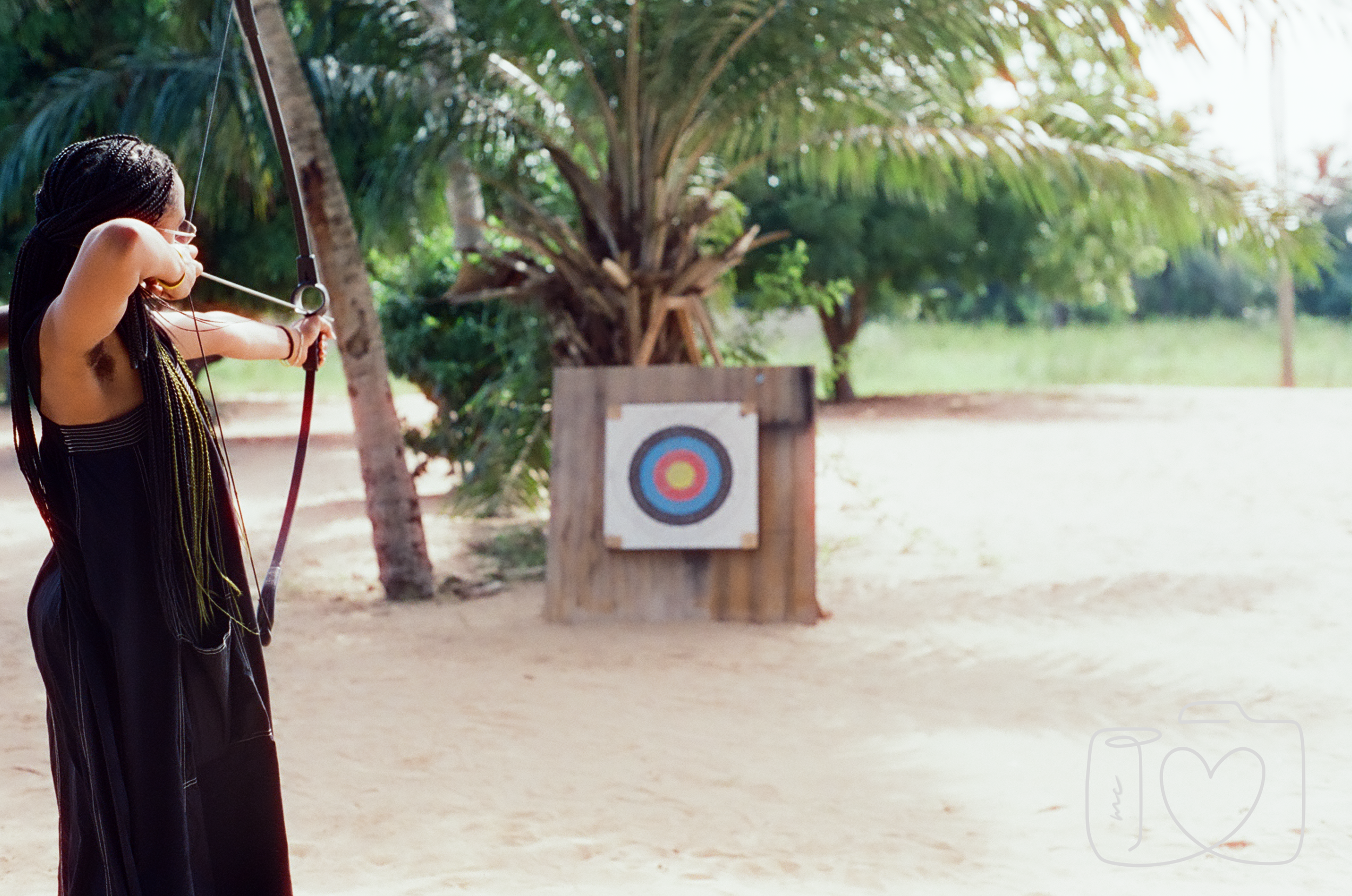 A girl with long braids practicing archery outdoors on a sandy area, aiming her bow at a target with colorful rings in front of trees and greenery.