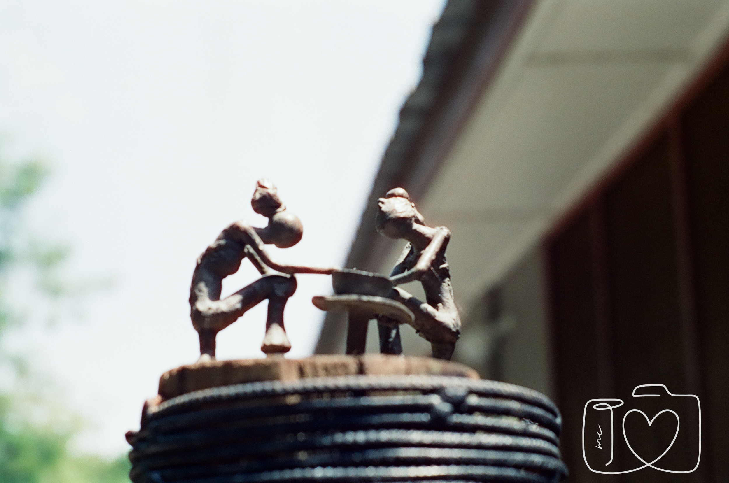 A metal sculpture of two people sitting at a table having a meal, placed on top of a stack of plates, with part of a house or building and sky in the background.