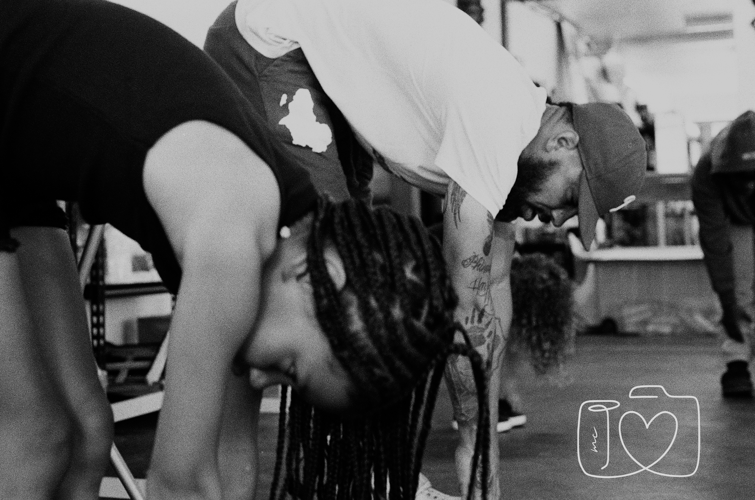 A person with braided hair and tattoos leaning forward in a yoga pose on a yoga mat, in a room with shelves and other people in the background.