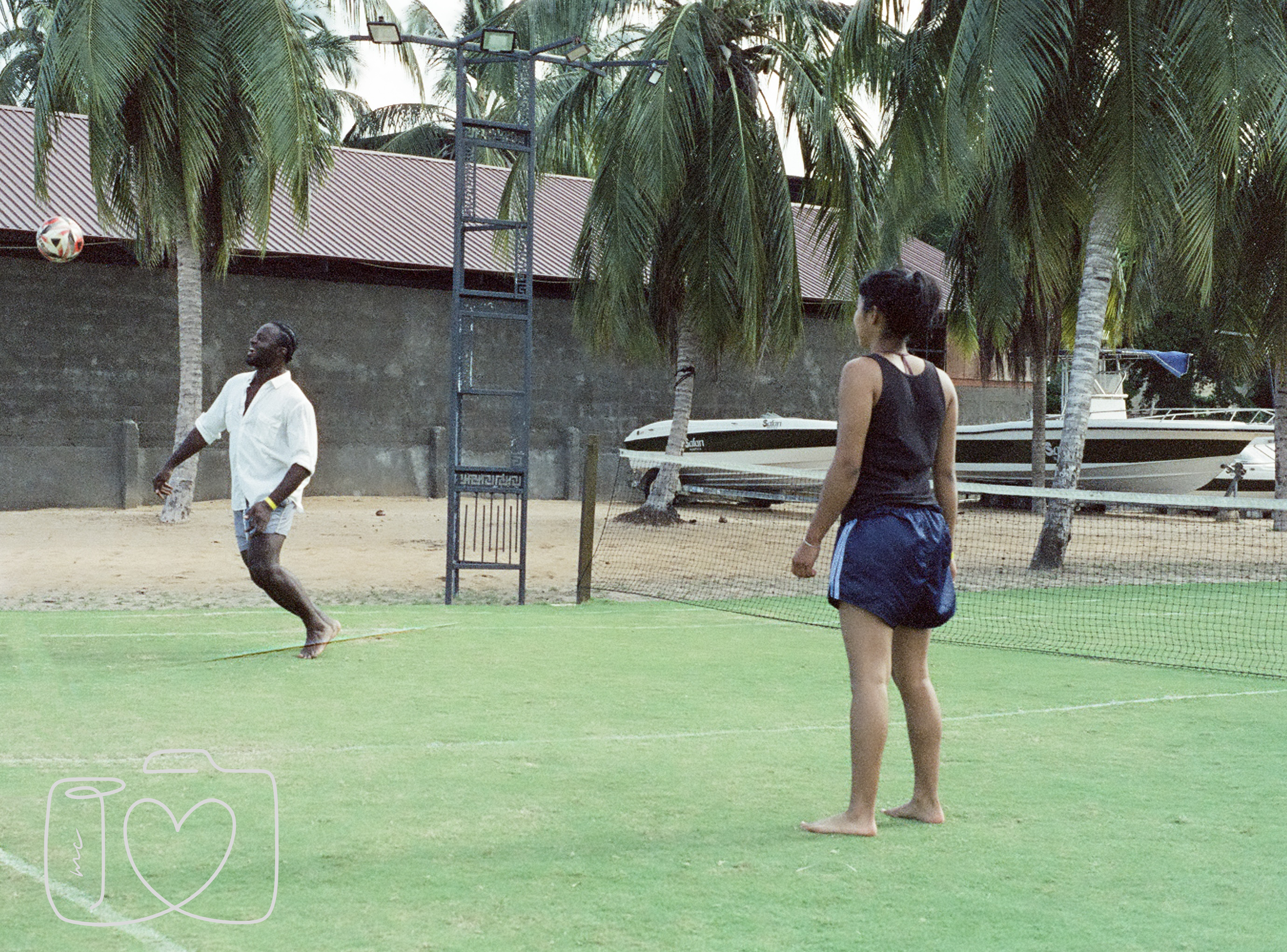 Two people playing volleyball on a grassy court with palm trees, boats, and a concrete wall in the background