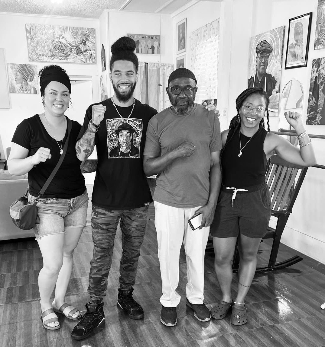 Four people standing together in a room, smiling and posing with fists raised. The room has artwork on the walls and a rocking chair.