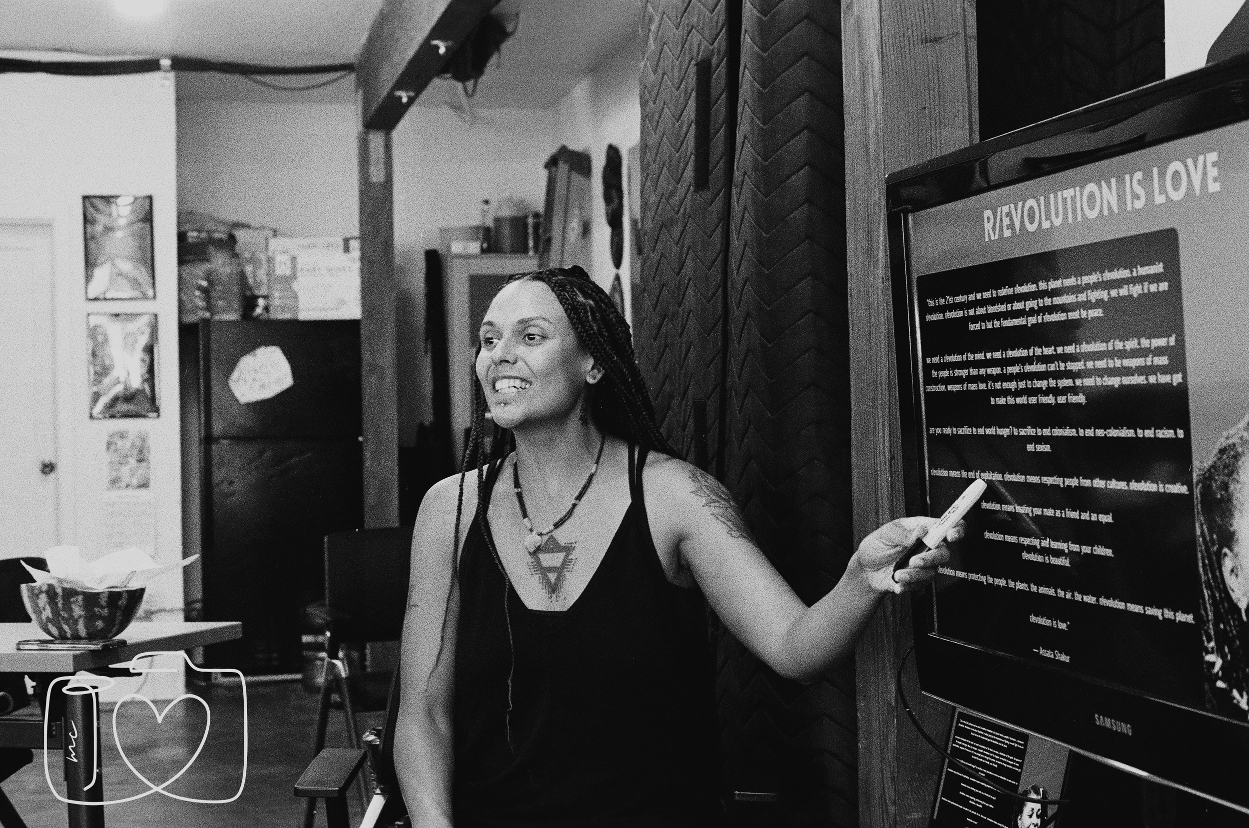 A woman with braided hair, tattoos, and a necklace, smiling and holding a remote control or pointer, standing next to a television screen displaying text titled 'Revolution is Love'. The room appears to be a casual or creative space with artwork on t