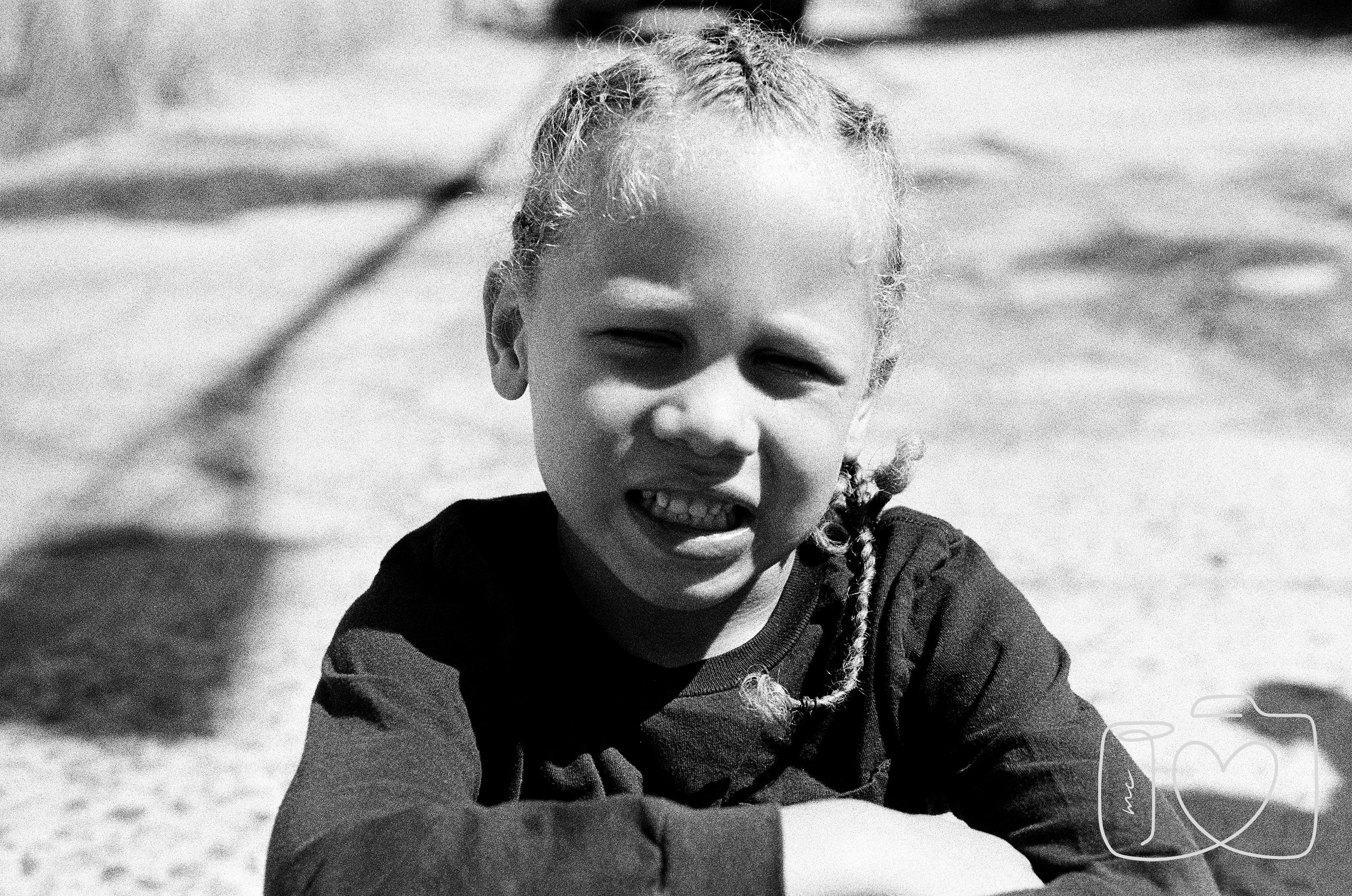 A young girl with curly hair styled in braids smiling and sitting outdoors in bright sunlight, with a blurred background.