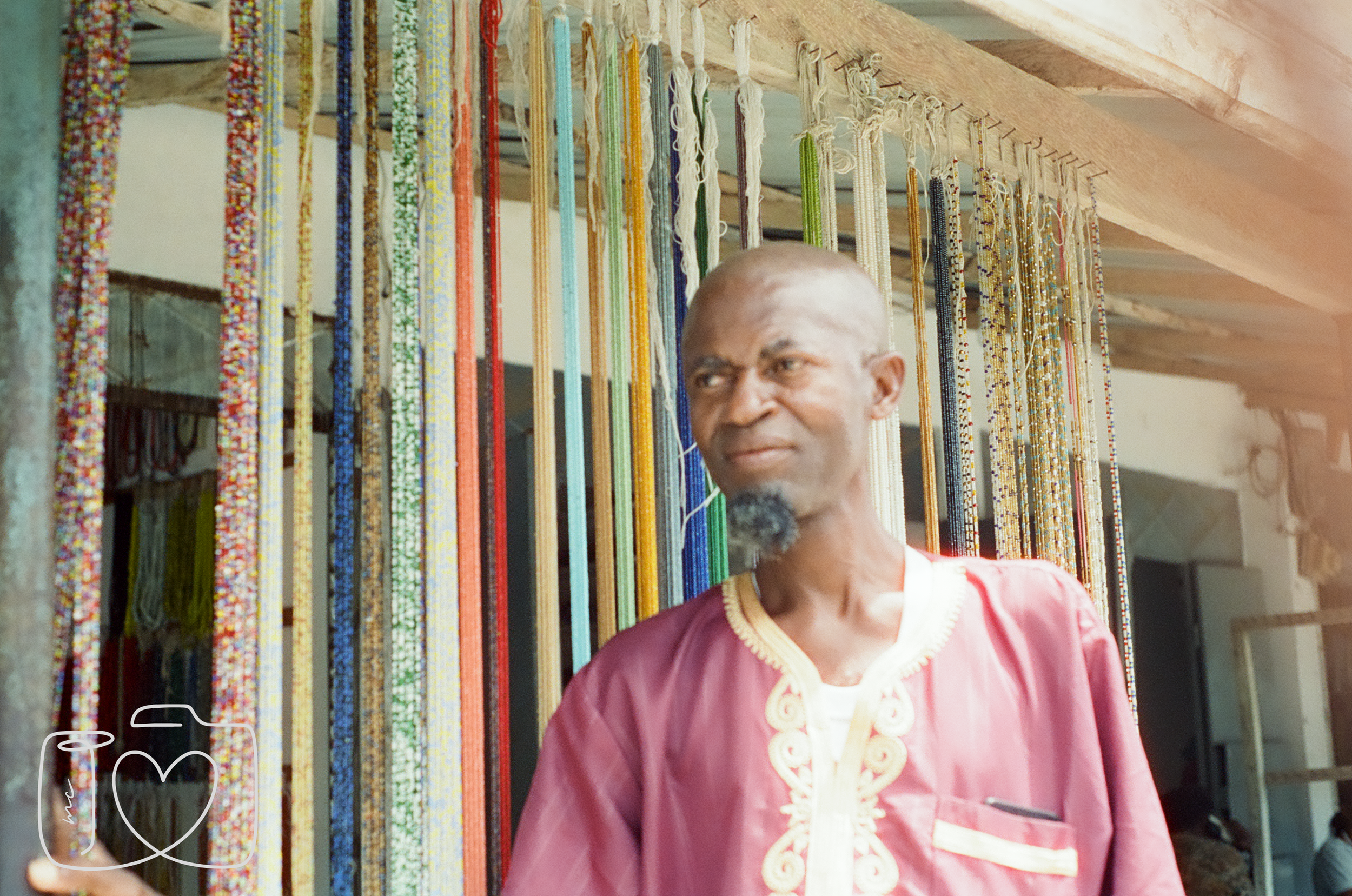 A man dressed in a pink traditional garment with gold embroidery stands in front of colorful strings of beads hanging in a shop.