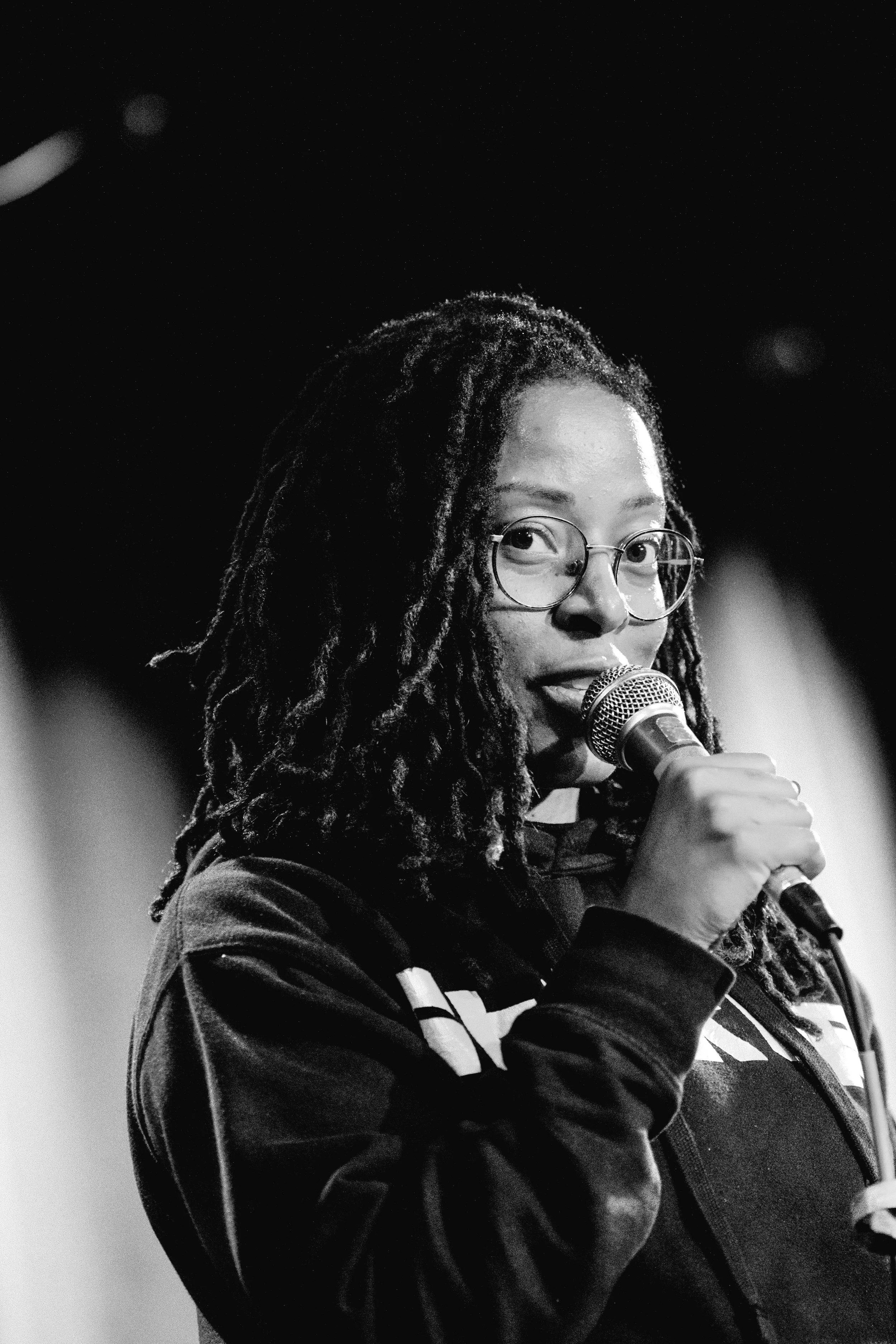 A woman with glasses and dreadlocks speaking into a microphone on stage.