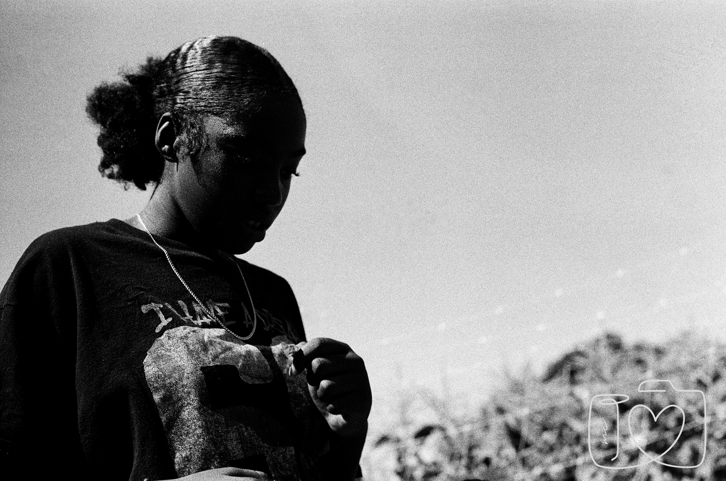 A black and white photo of a person with braided hair, wearing a T-shirt and a necklace, standing outdoors with rocky terrain in the background.