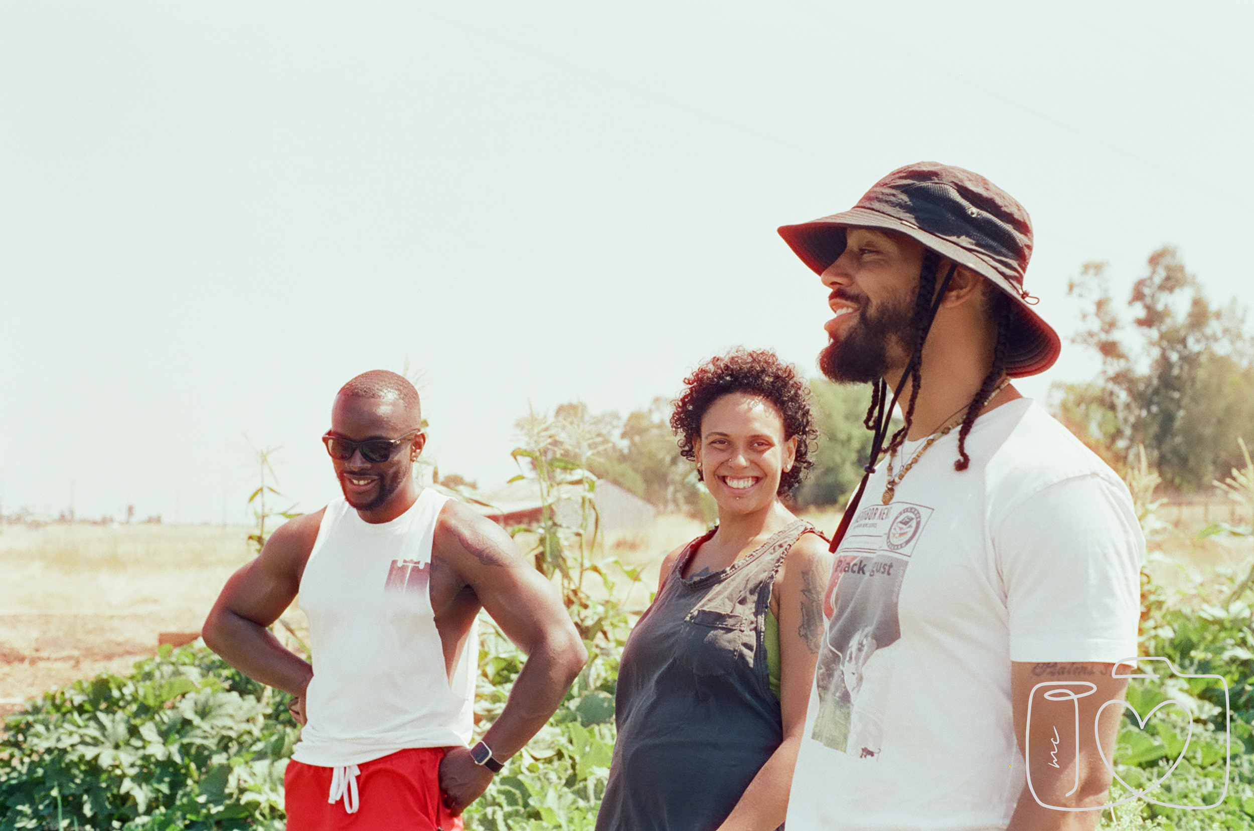 Three friends smiling outdoors on a sunny day in a field with green plants. The man on the left is wearing sunglasses and a white sleeveless shirt, the woman in the middle has curly hair and is wearing a black sleeveless top, and the man on the right