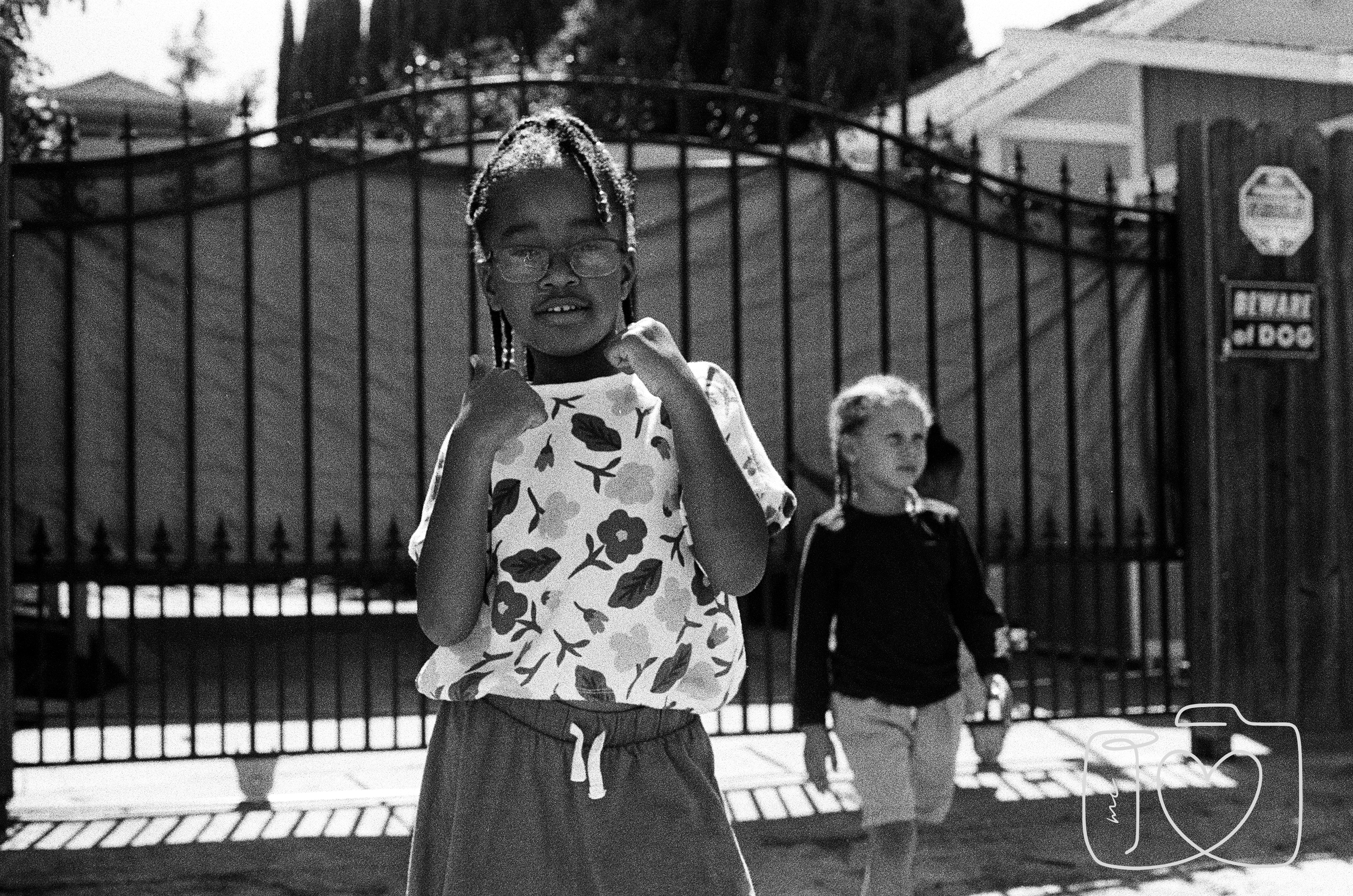 A young girl wearing glasses, a floral T-shirt, and shorts making a fist with both hands, standing outdoors in front of a gate, with another girl in the background near the gate.