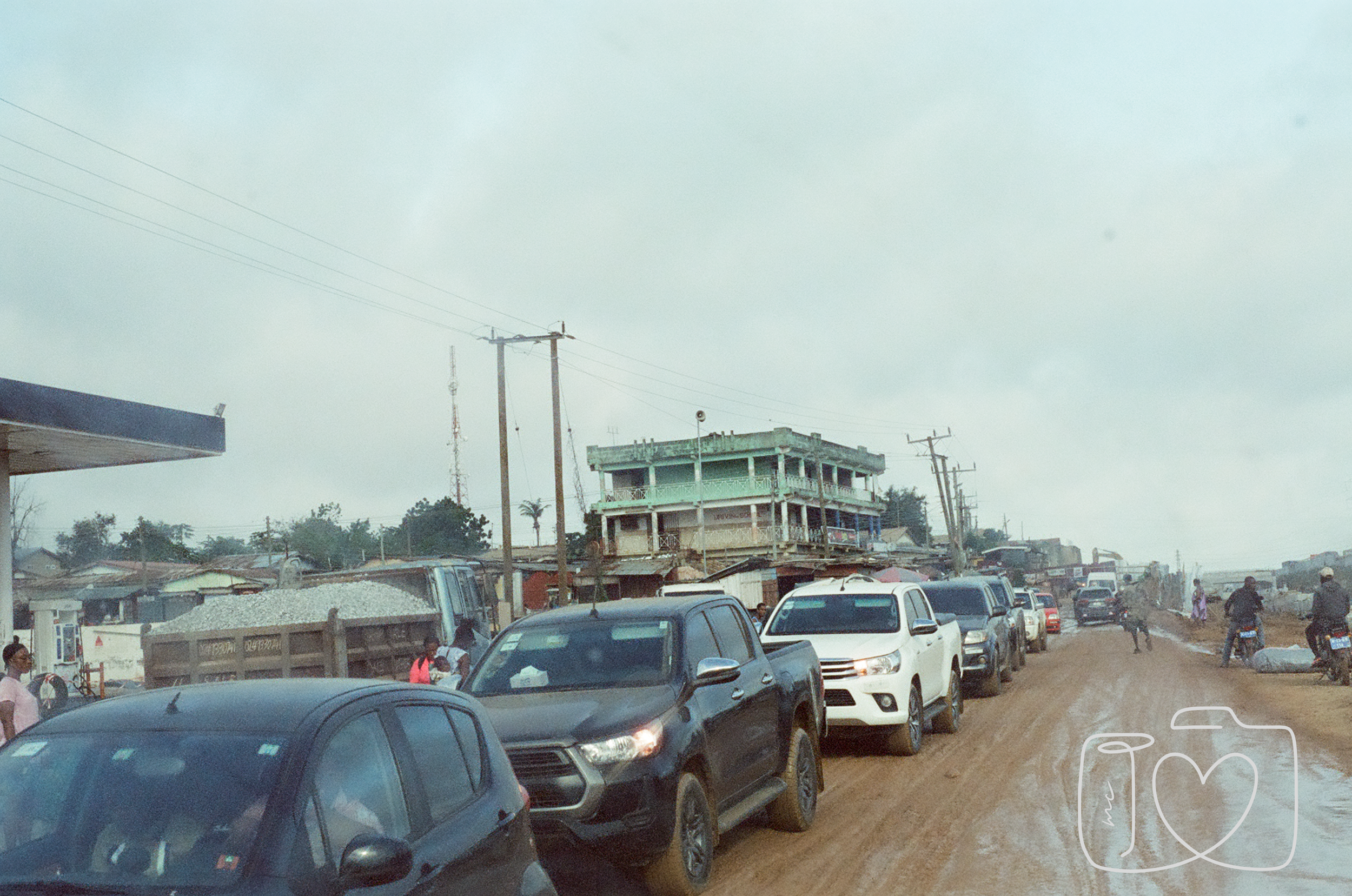 A busy dirt road with multiple cars and motorcycles, in a small town with simple buildings and utility poles under a cloudy sky.