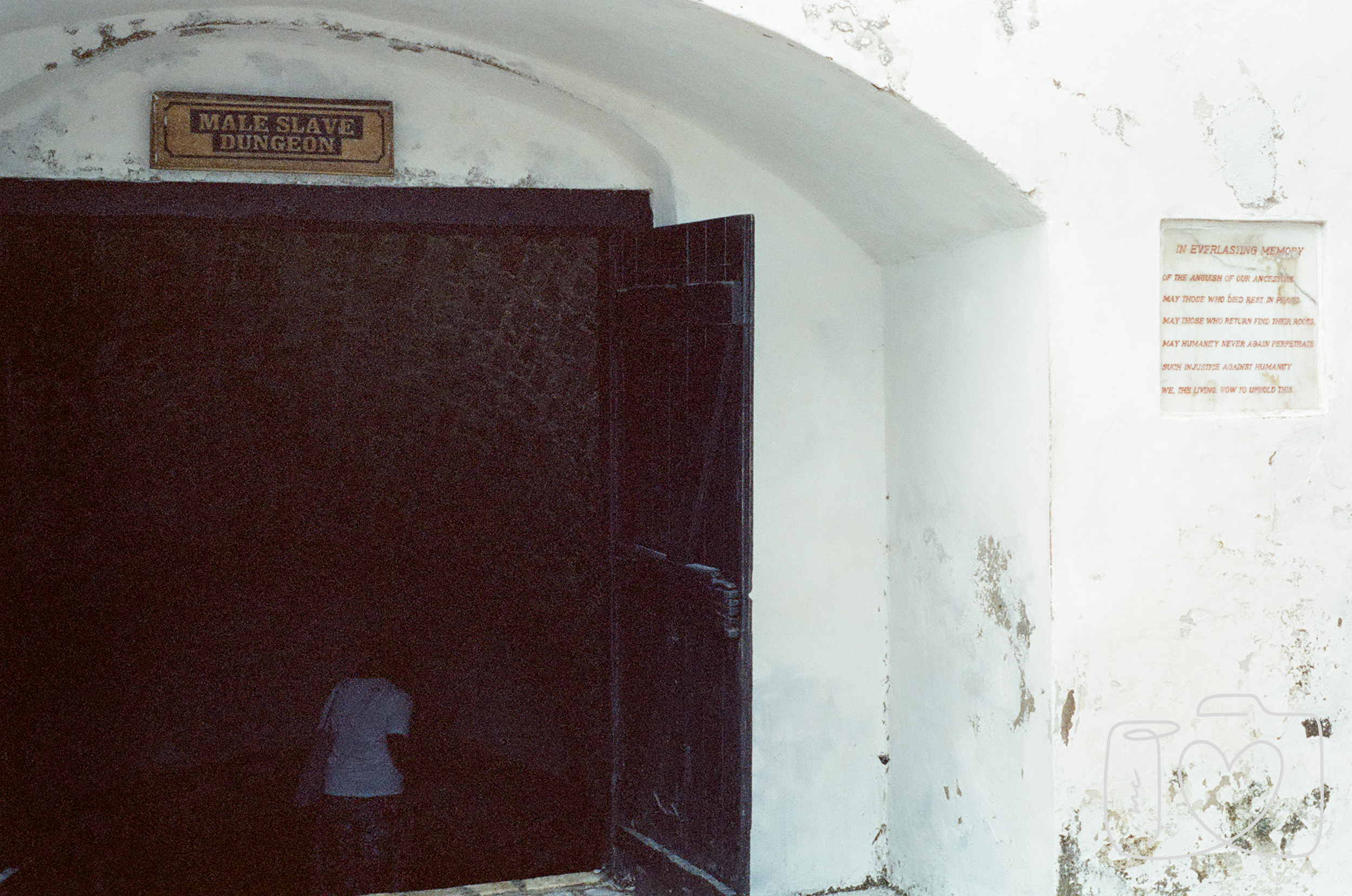 A dark doorway with a sign above it labeled 'Male Slave Dungeon'. Inside the darkness, a person is faintly visible, wearing a white shirt and dark pants. To the right of the door, there is a white wall with peeling paint and a plaque with red text, p