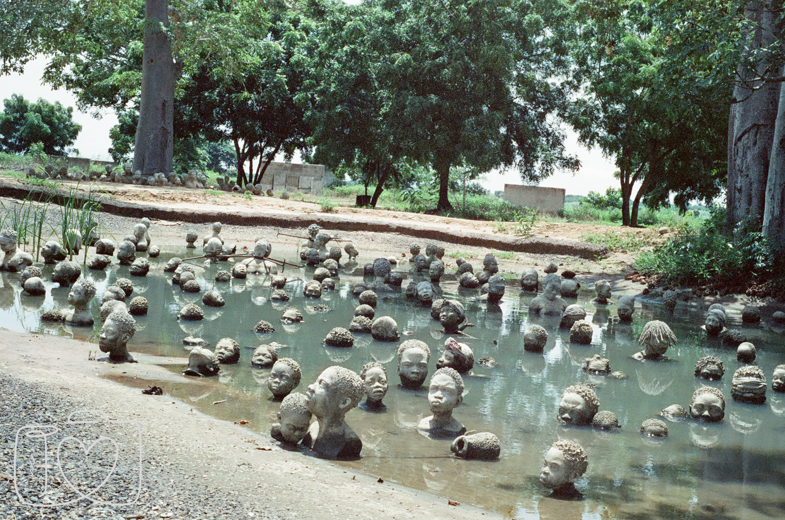 A pond filled with numerous small head sculptures emerging from the water, surrounded by trees and greenery under a bright sky.