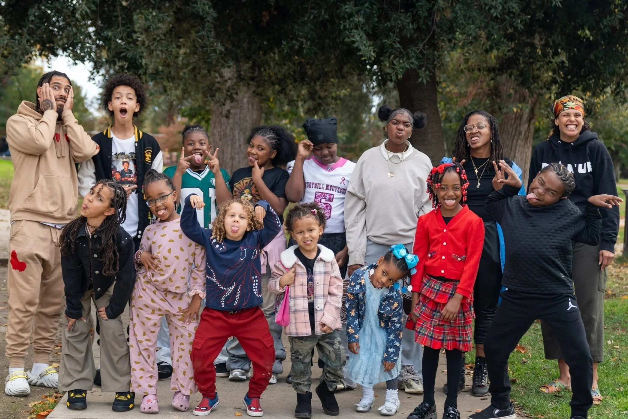 A group of children and adults posing outdoors, making funny faces and smiles, with trees in the background.