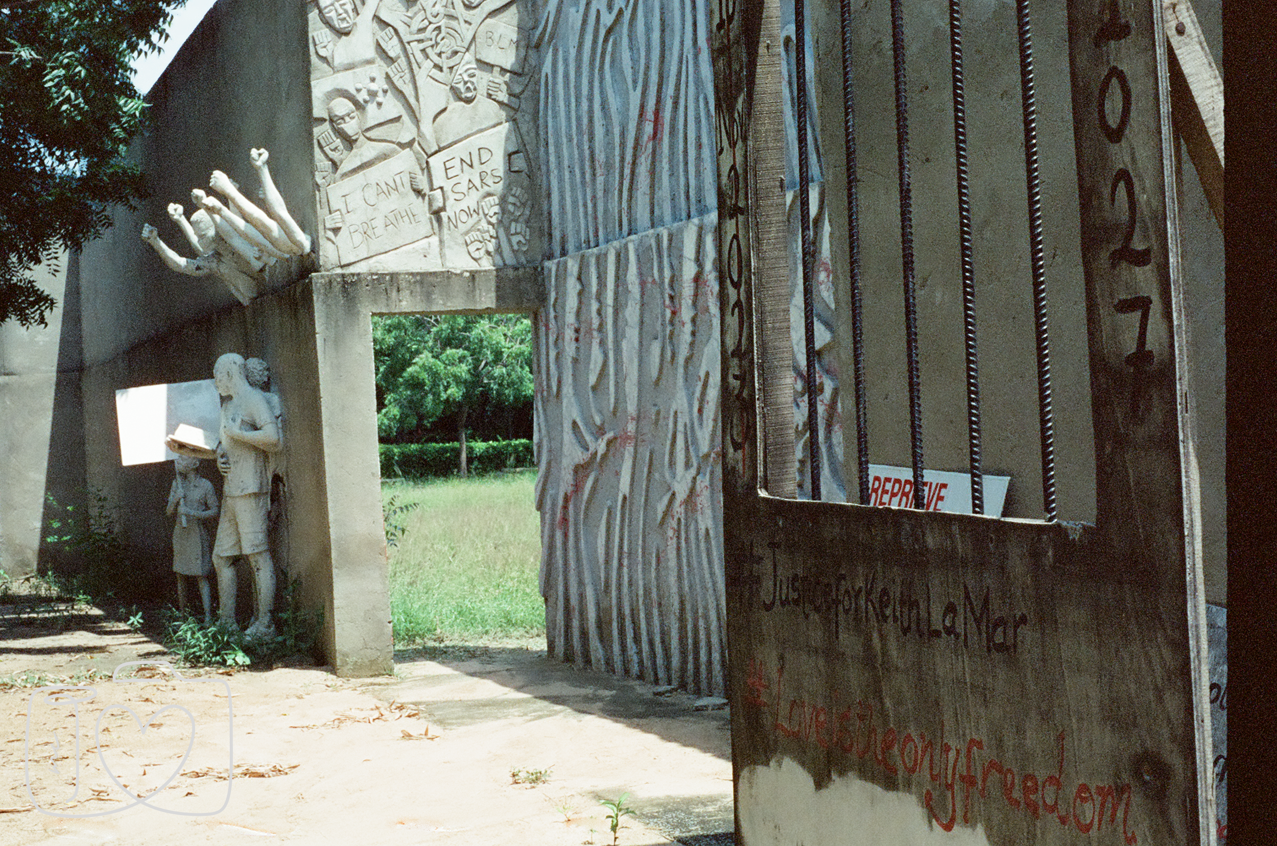 A concrete sculpture featuring human figures and grass with trees in the background, a parapet with barred windows, and signs including one that says 'REPREVE' and another with a website address 'www.loveconsciousfreed.com'.