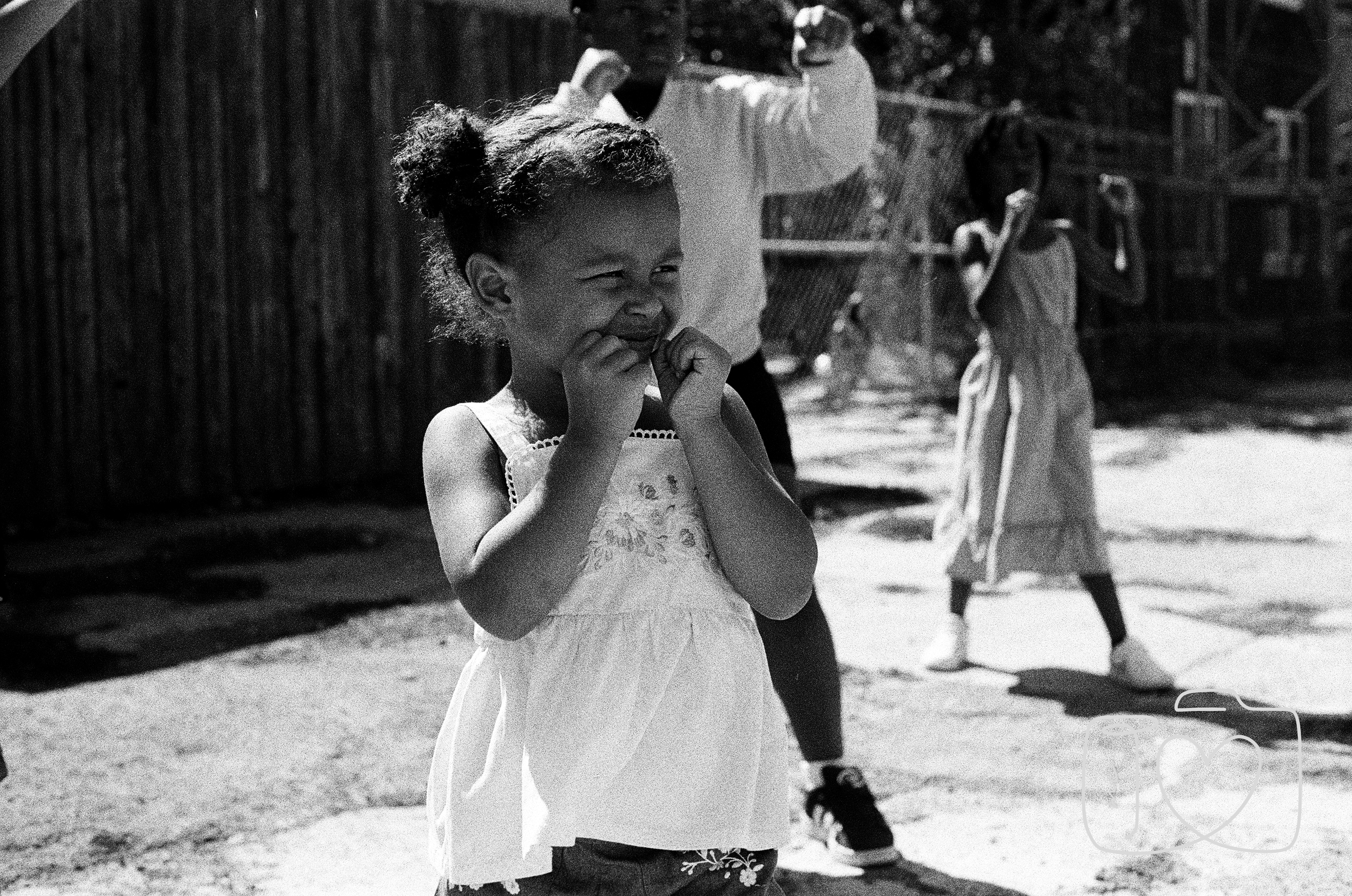 Smiling young girl in a light-colored dress with floral embroidery, standing outdoors with her hands near her face, and other children playing in the background.