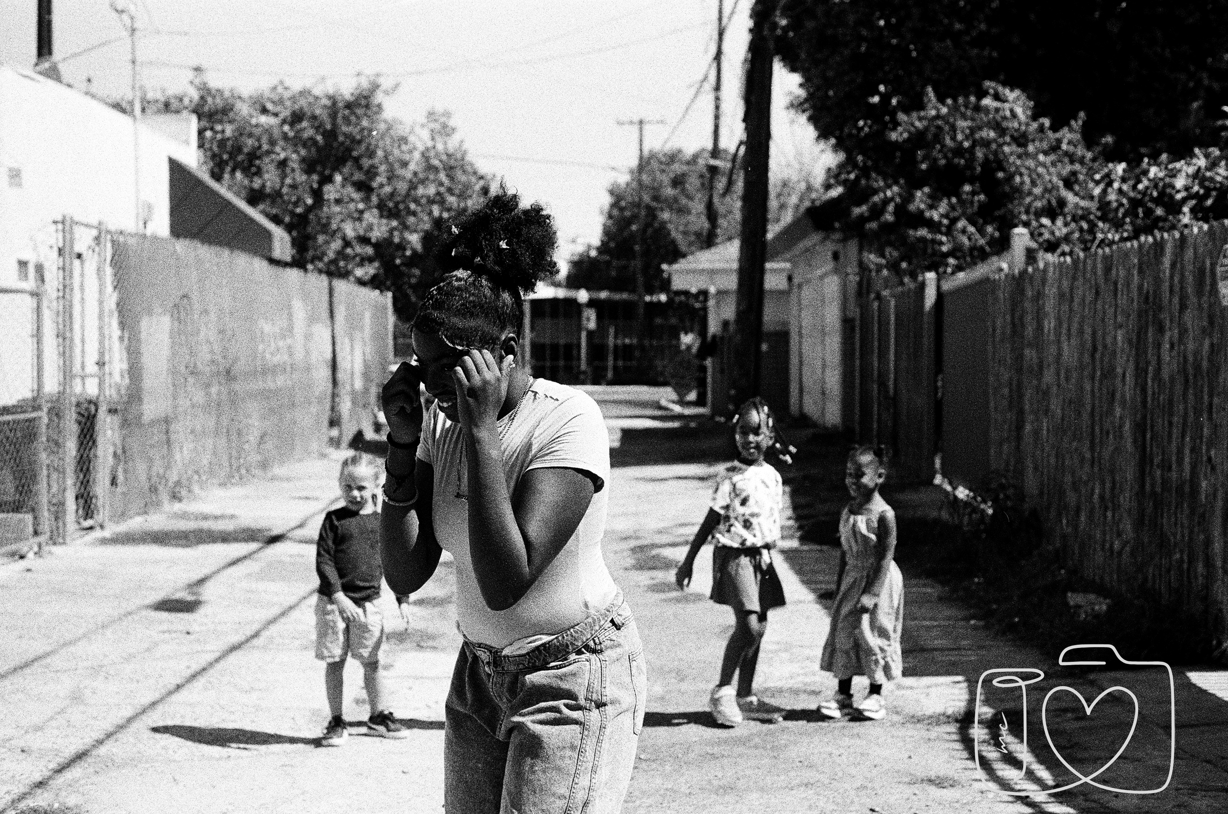 A young woman with curly hair in a ponytail talking on a cell phone in an alleyway with three children in the background, two girls and a boy, standing near a fence and trees.