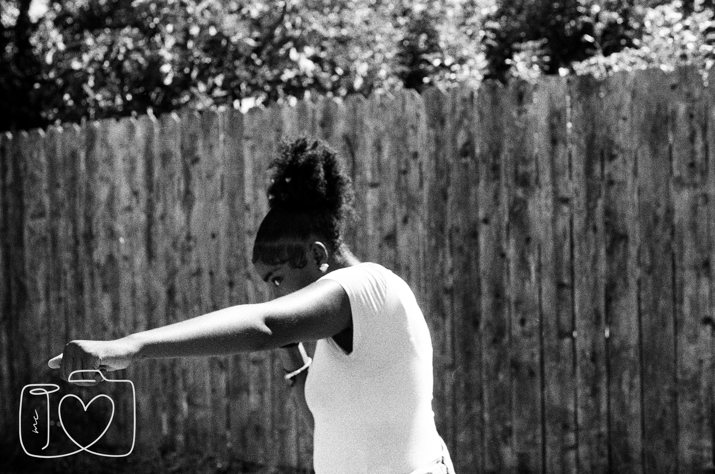 A woman with curly hair tied up in a bun is standing outdoors, looking downward with her arms extended to the side, wearing a white T-shirt, in front of a wooden fence with trees and foliage in the background.