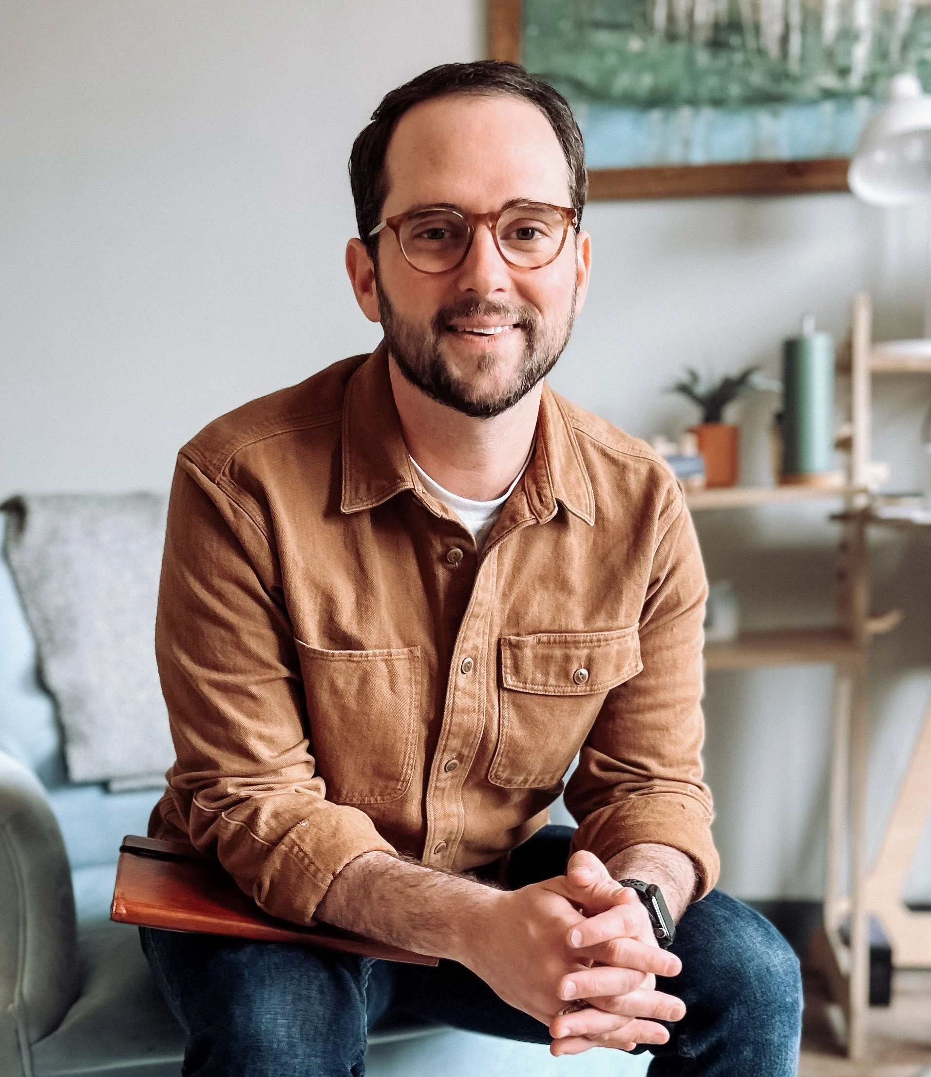 A man with brown hair and glasses sitting on a gray couch in a living room, smiling, wearing a brown jacket, holding a folder.